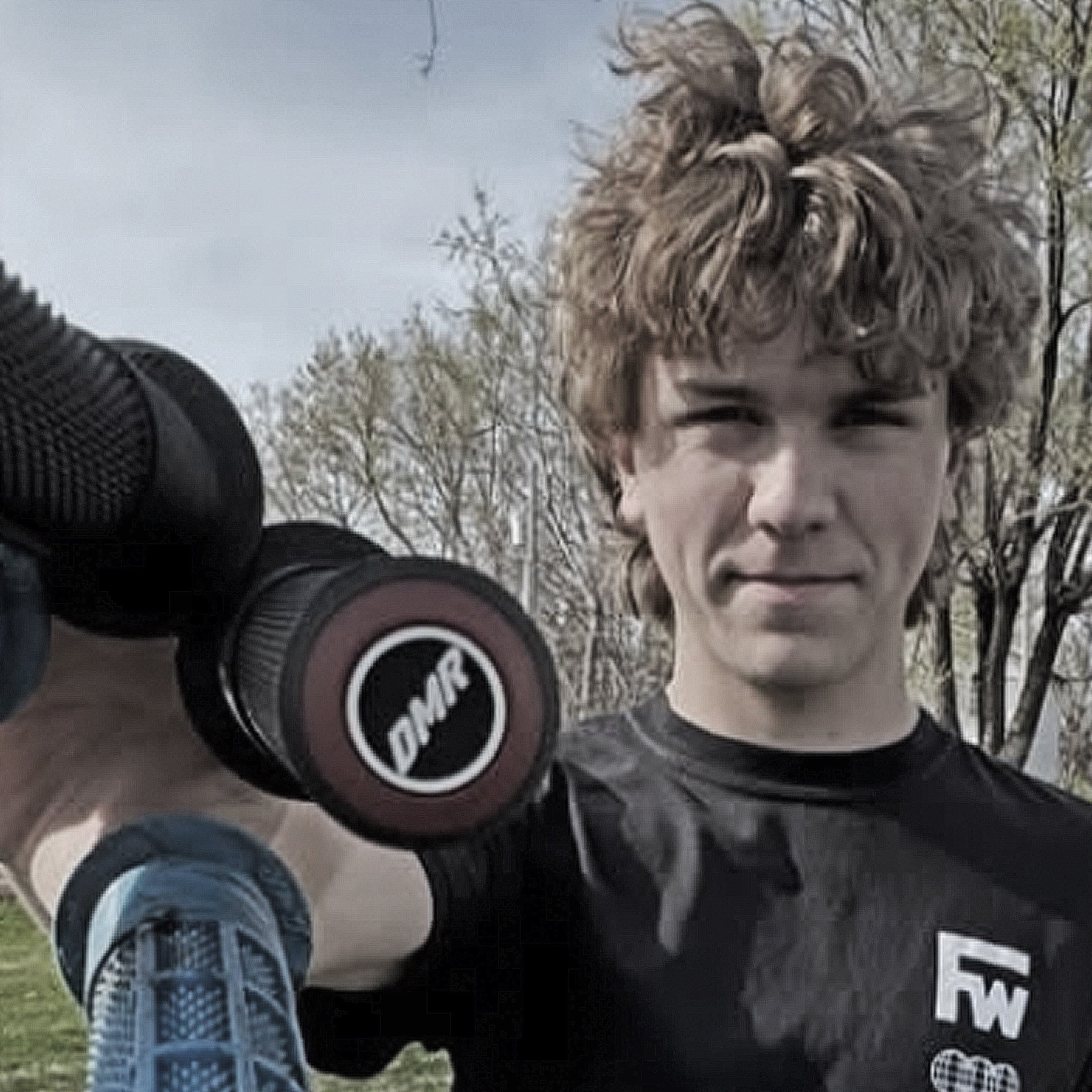 Person holding a camera with a lens, wearing a black shirt with visible branding, outdoors.