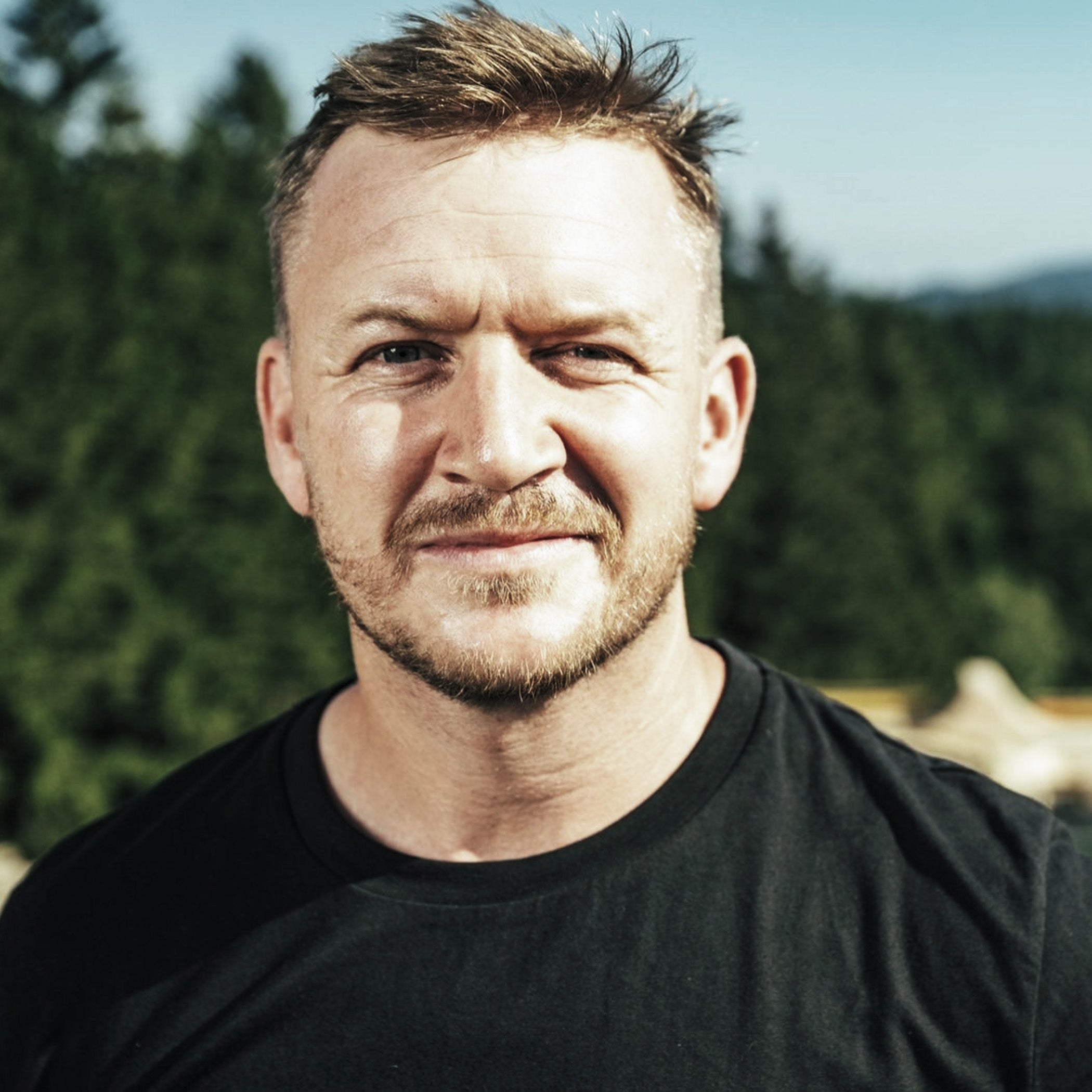 A close-up portrait of a man with short light brown hair and a light beard, wearing a black crew-neck t-shirt, set against a blurred background of green trees and a blue sky.