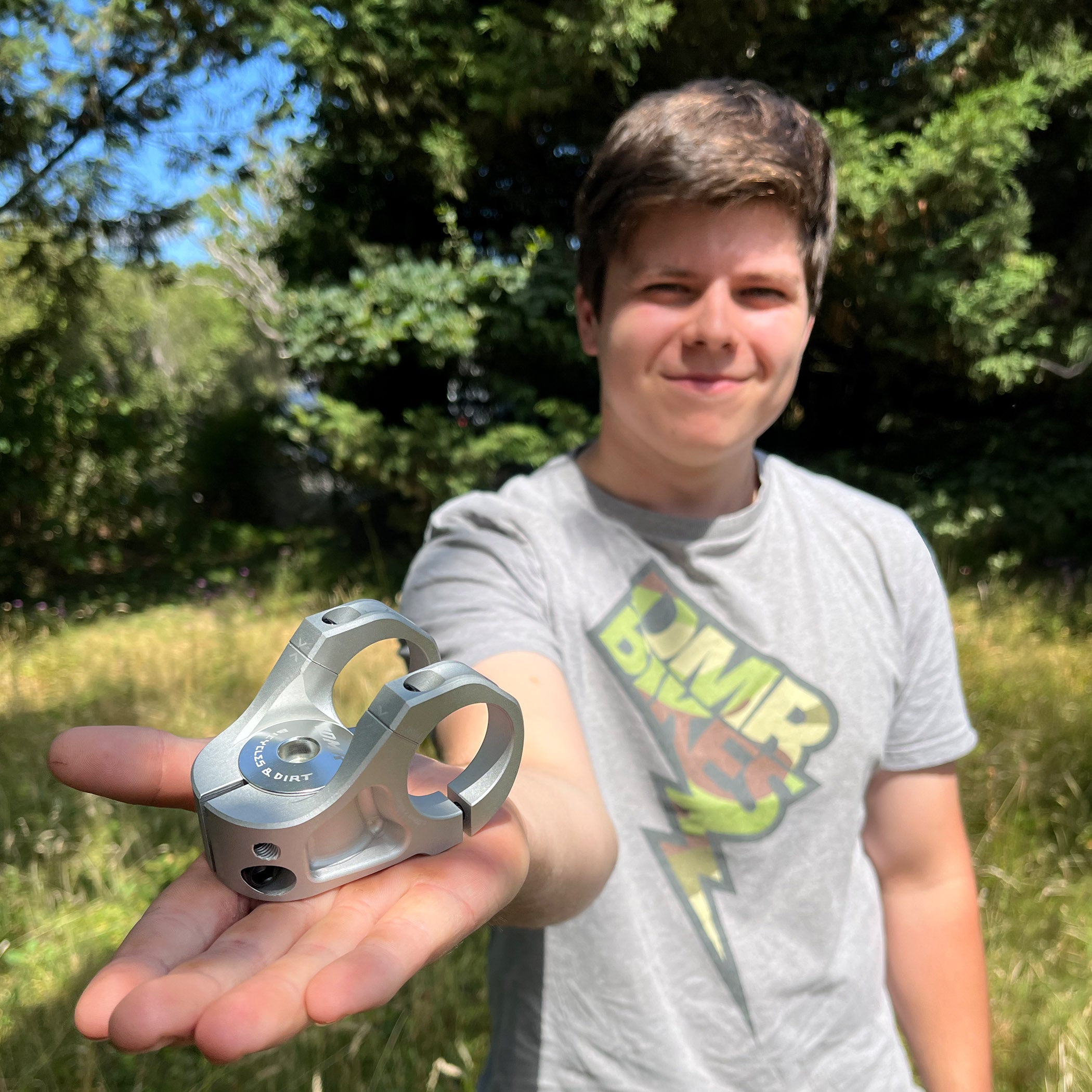 DMR team rider holding up a silver metallic bicycle stem in his hand, with trees and grass in the background.