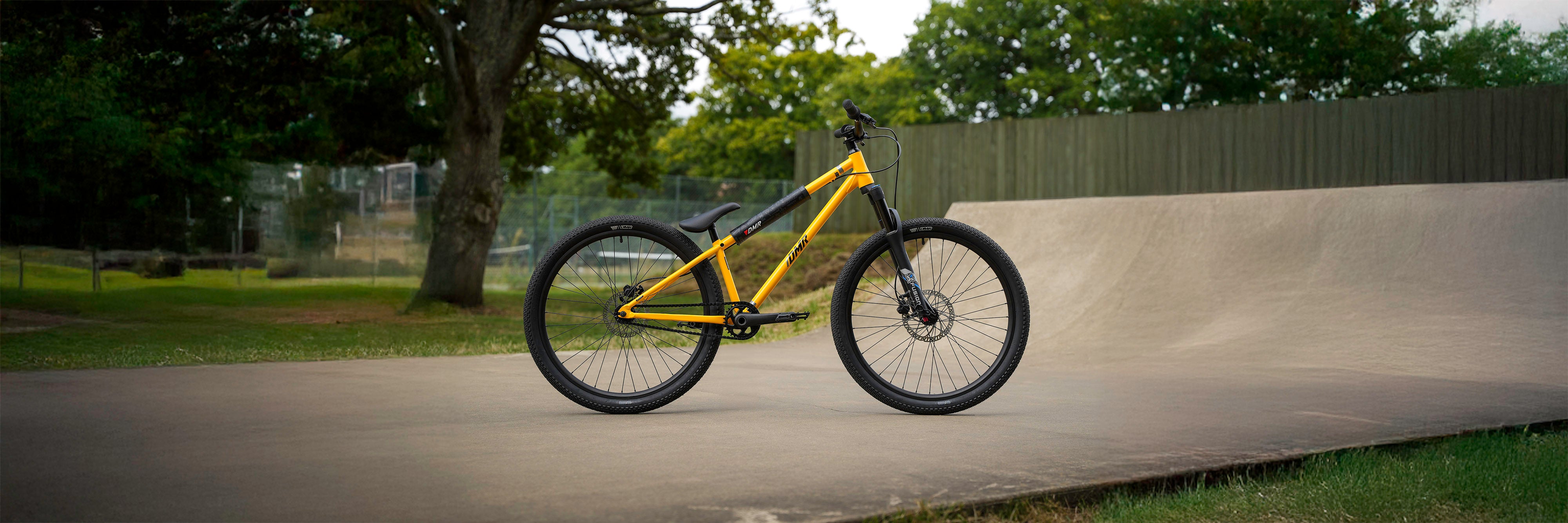 DMR Sect Pro dirt jumping jumper bike bicycle bicycling product, in Yellow, placed upon a skate park ramp, a tree, fence and tennis court can be seen in the background.