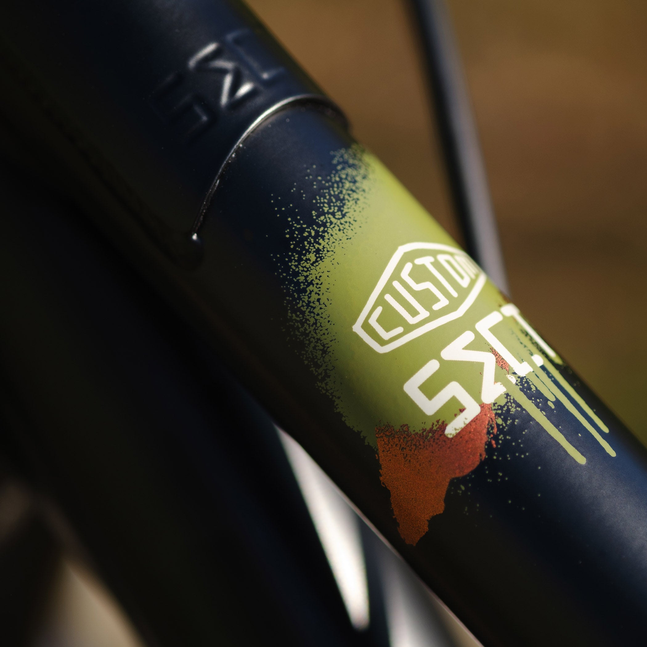 Close-up of a DMR Sect Custom 24" off-road mountain cycling cycle bike bicycle frame's top tube, in matt black, with visible branding. A blurred brown background can be seen.