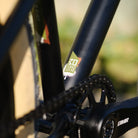 Close-up of a DMR Sect Custom 24" off-road mountain cycling cycle bike bicycle frame's down tube and bottom bracket area, in matt black, with visible branding. A blurred brown background can be seen.