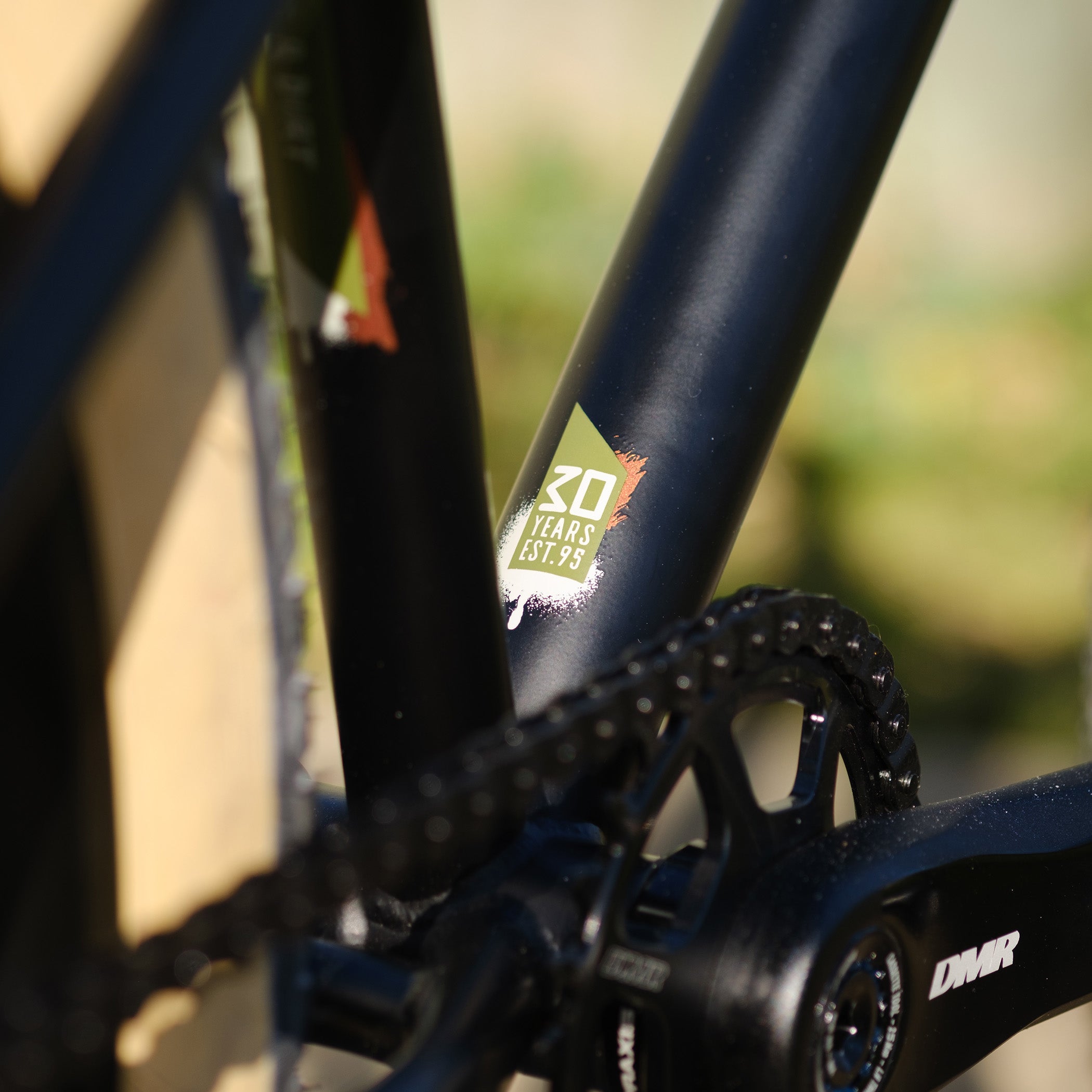 Close-up of a DMR Sect Custom 24" off-road mountain cycling cycle bike bicycle frame's down tube and bottom bracket area, in matt black, with visible branding. A blurred brown background can be seen.