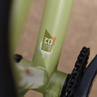 Close-up of a DMR Sect Custom 26" off-road mountain cycling cycle bike bicycle frame's down tube and bottom bracket area, in pistachio green, with visible branding. A blurred brown background can be seen.