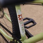 Close-up of a DMR Sect Custom 26" off-road mountain cycling cycle bike bicycle frame's seat tube, in pistachio green, with visible branding. A blurred brown background can be seen.