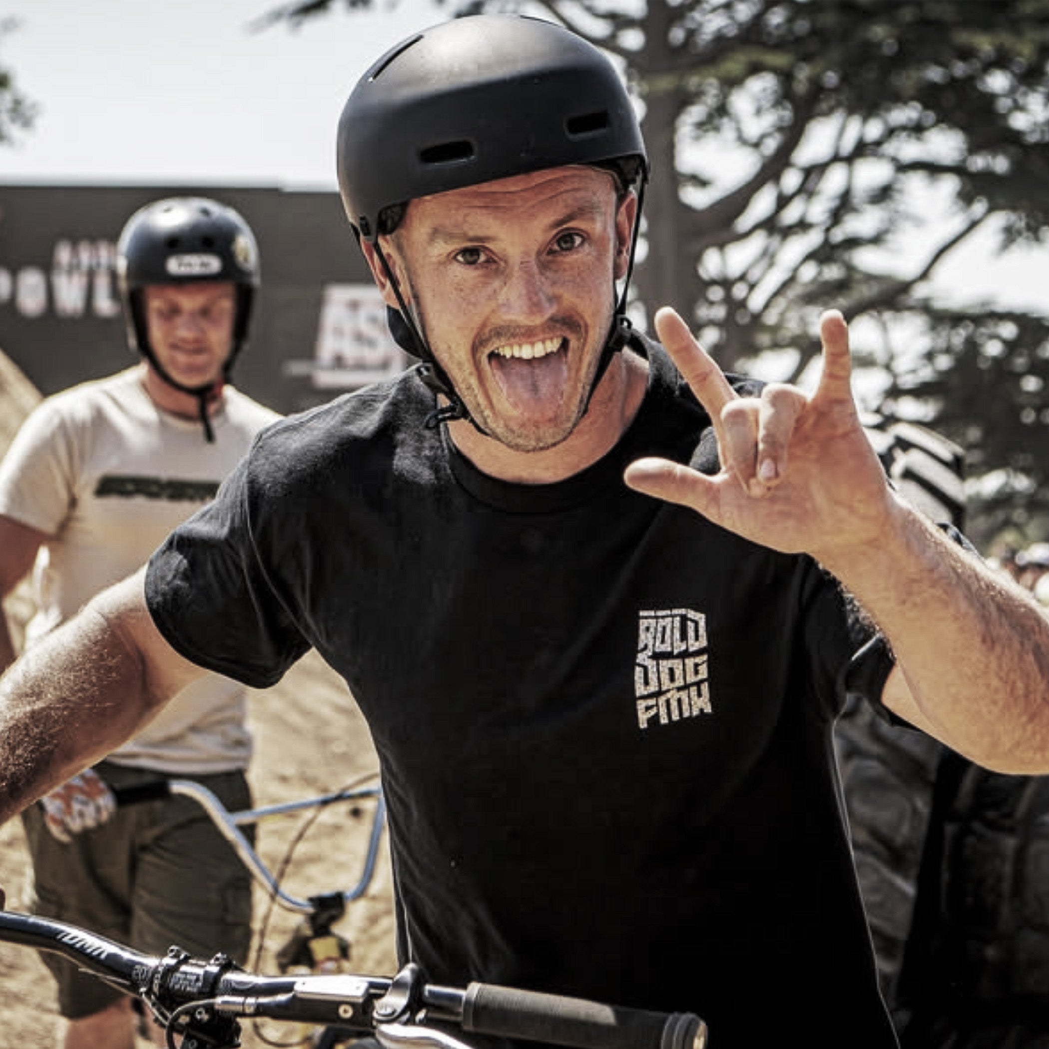 A man wearing a black helmet and a black t-shirt with a gold 'BOLD DOG FMX' logo on the chest, making a rock on gesture with his hand while riding a bicycle outdoors. Another person is visible in the background.