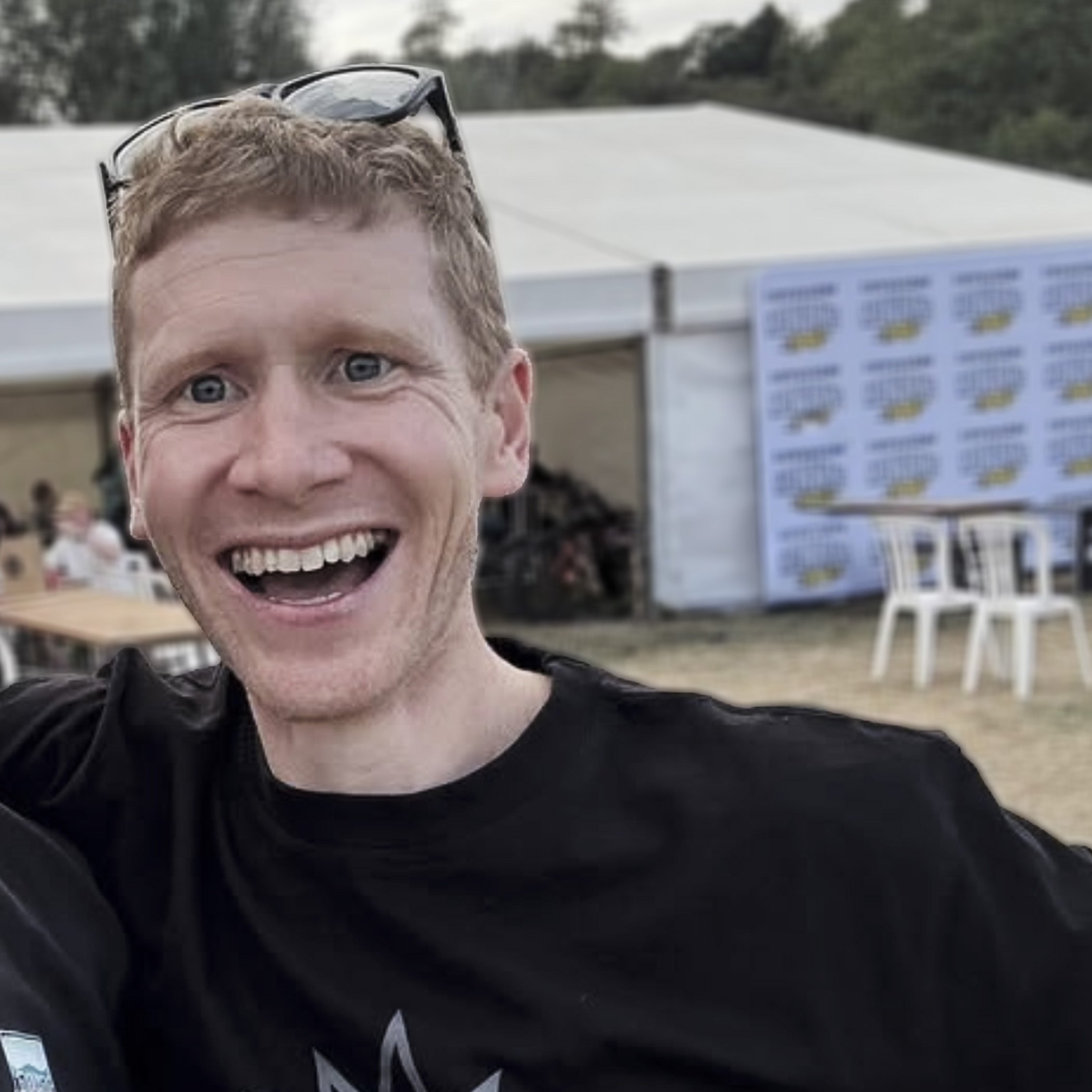 A smiling man with short blond hair and sunglasses on his head wearing a black t-shirt, photographed in an outdoor event setting with a white tent and banner in the background.