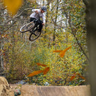 A person with a name of Andy Lehmann, riding on a DMR Rhythm off-road mountain cycling bicycle bike, performing an aeronautical manoeuvre in a forest with autumn leaves.