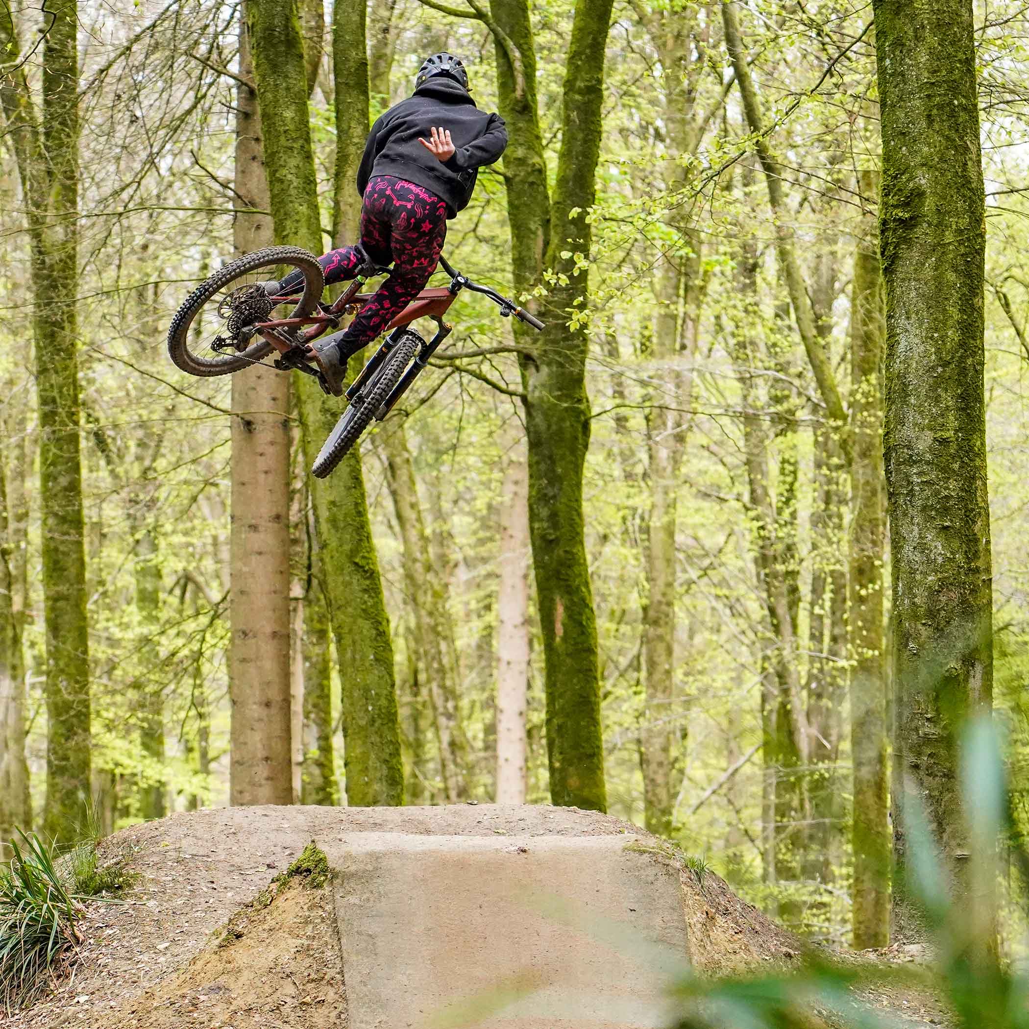 A person by the name of Preston Williams, riding on an off-road mountain cycling bicycle bike, performing an aeronautical manoeuvre requiring removal of one of his hands in the air, over a jump in an open woodland.