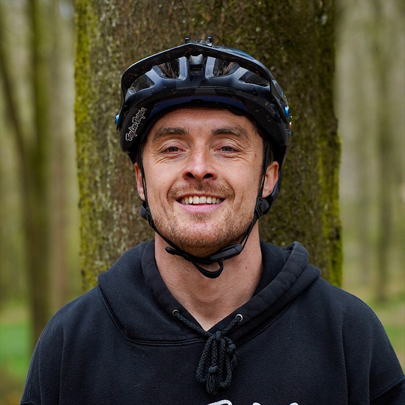 A portrait photograph of a person using the name Olly Wilkins, stood in front of a moss-covered tree, trying his best to look natural and smiling at the camera.