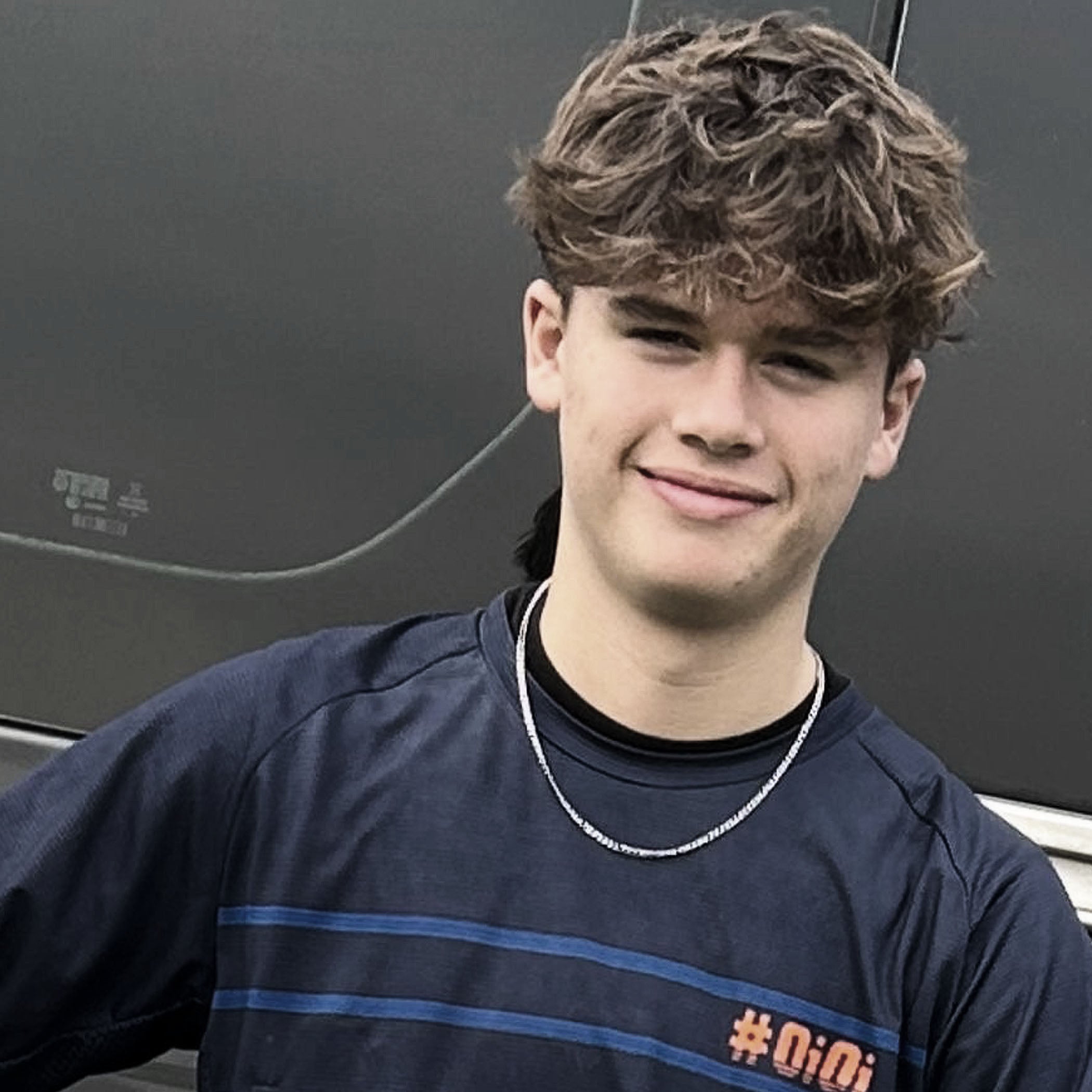 Young man with curly hair wearing a dark blue shirt with a chain necklace, standing against a dark background.