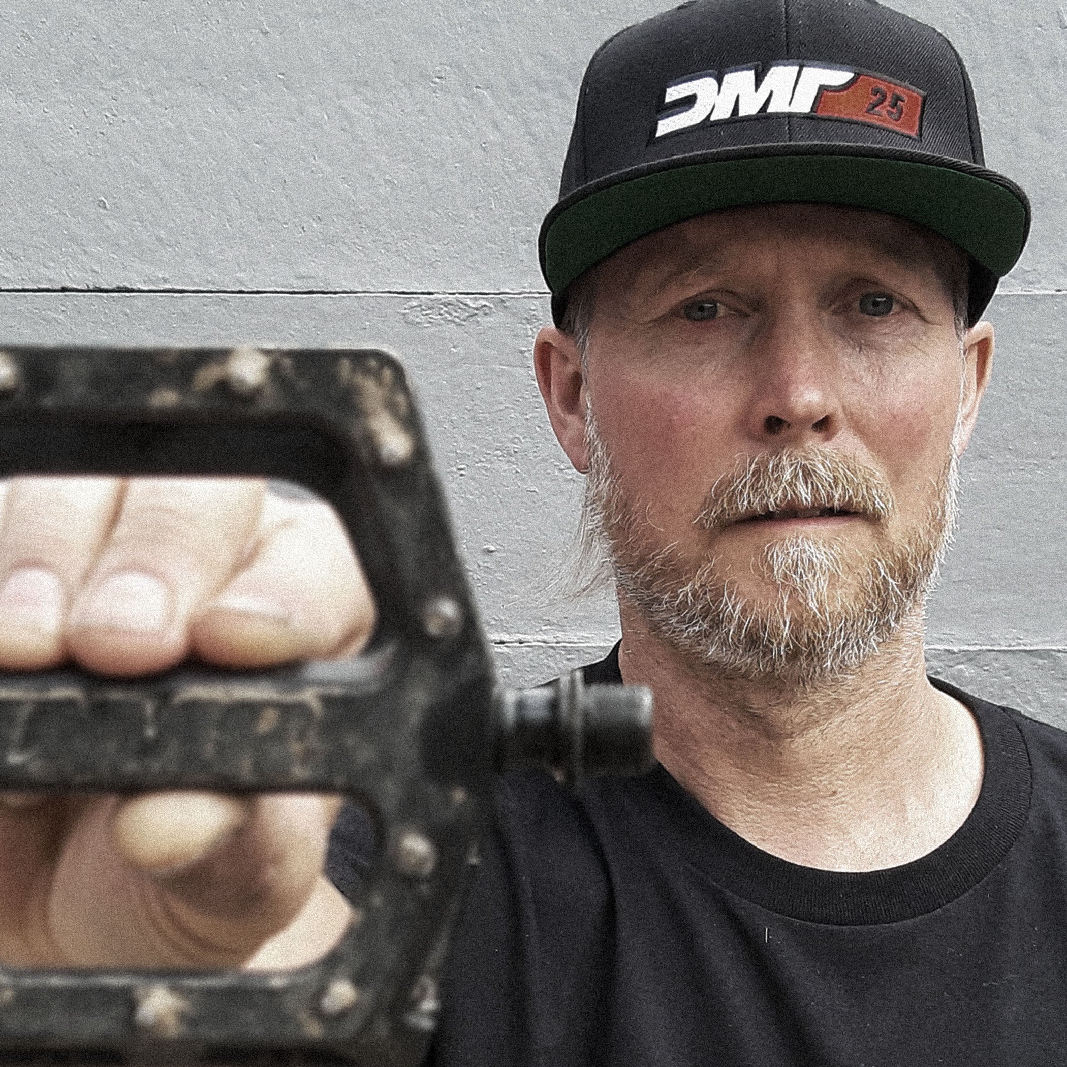 really old man wearing a cap with a logo, holding a tool against a gray background