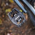 Black DMR VMAX off-road mountain cycle cycling pedal pedalling product, on a blurry brown background, featuring leaves and pine needles.