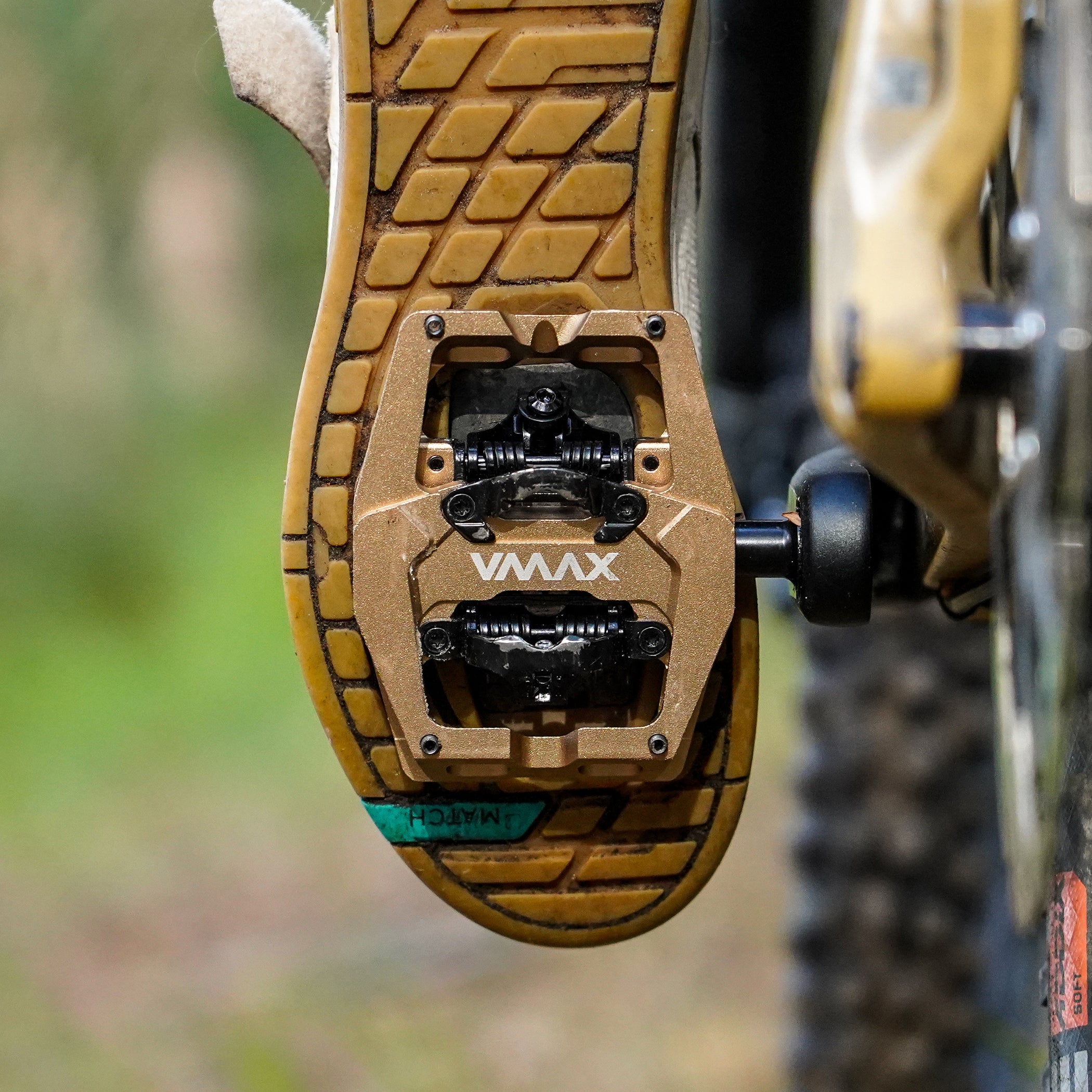 Bronze DMR VMAX off-road mountain cycle cycling pedal pedalling product, under foot of a human rider. The background is blurry and green, with brown elements.
