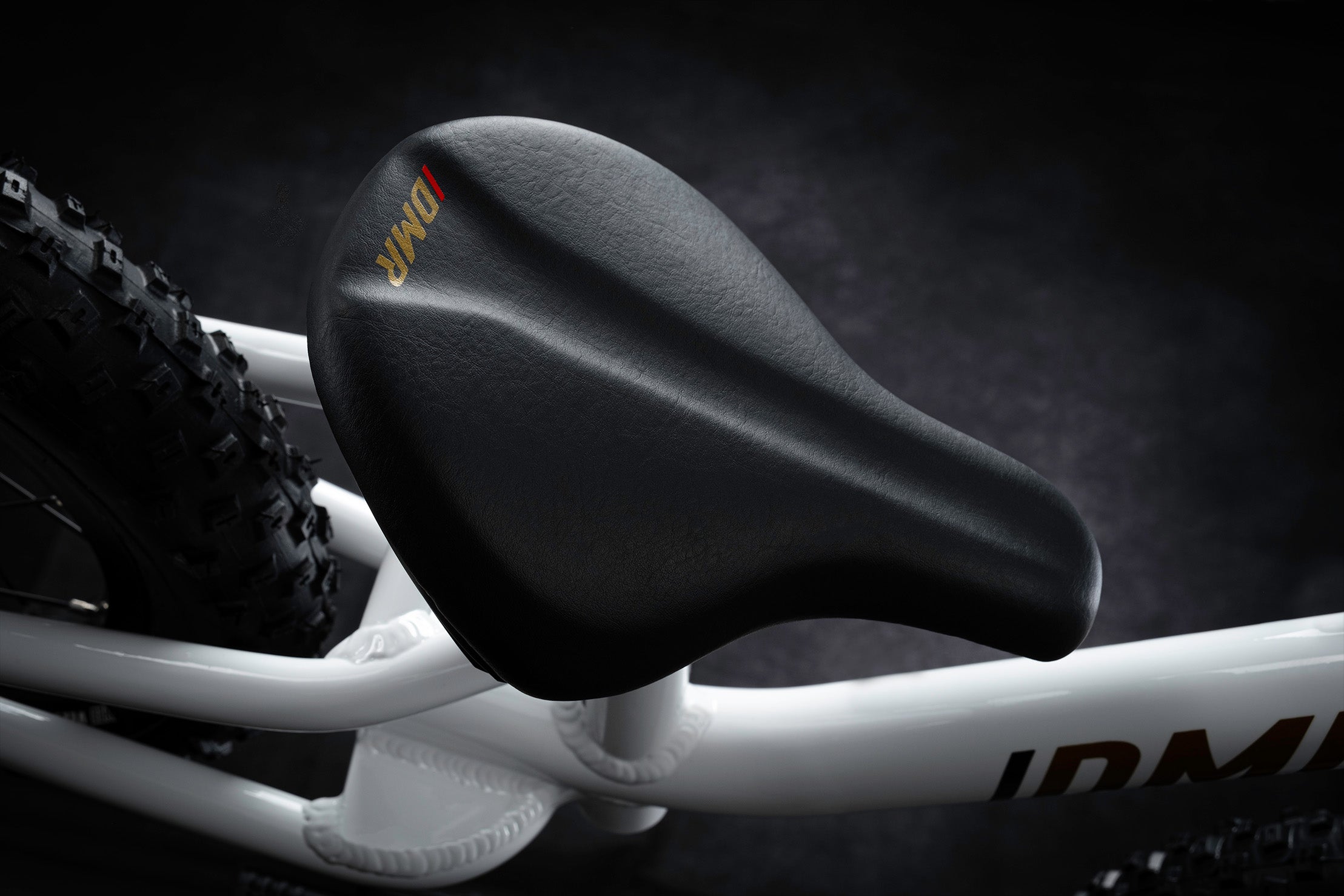 close-up of the dmr sidekick balance bicycle kids childrens bike saddle seat, with gold and red coloured dmr bicycles and dirt logo, the background is grey and black.
