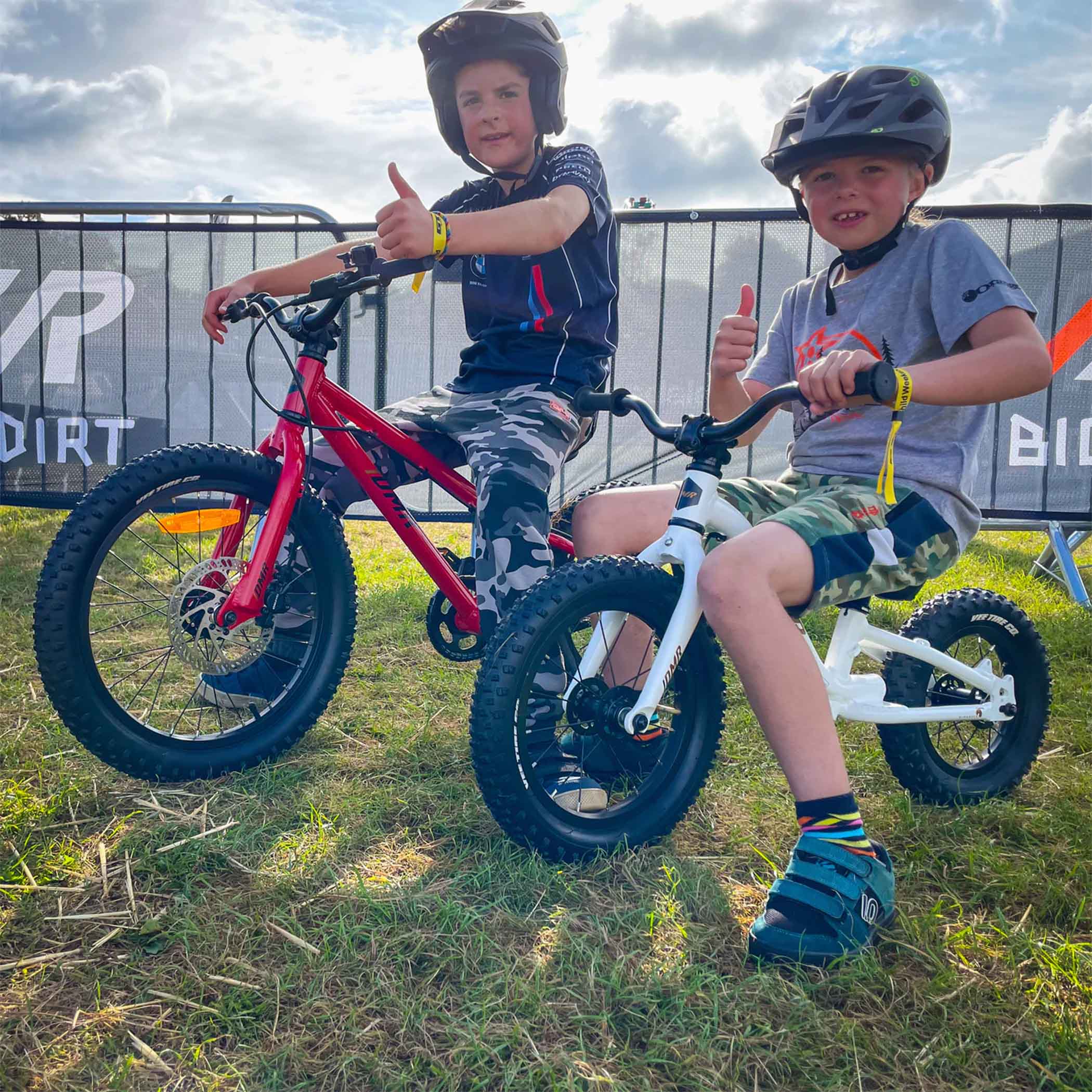 Two children, one sitting upon a dmr sidekick balance bike bicycle, the other sitting upon a dmr sidekick ride bicycle kids bike, each child is giving the thumbs up emoji in real life, whilst situated in a grassy area in front of a metal barrier fence, with a cloudy sky overhead.