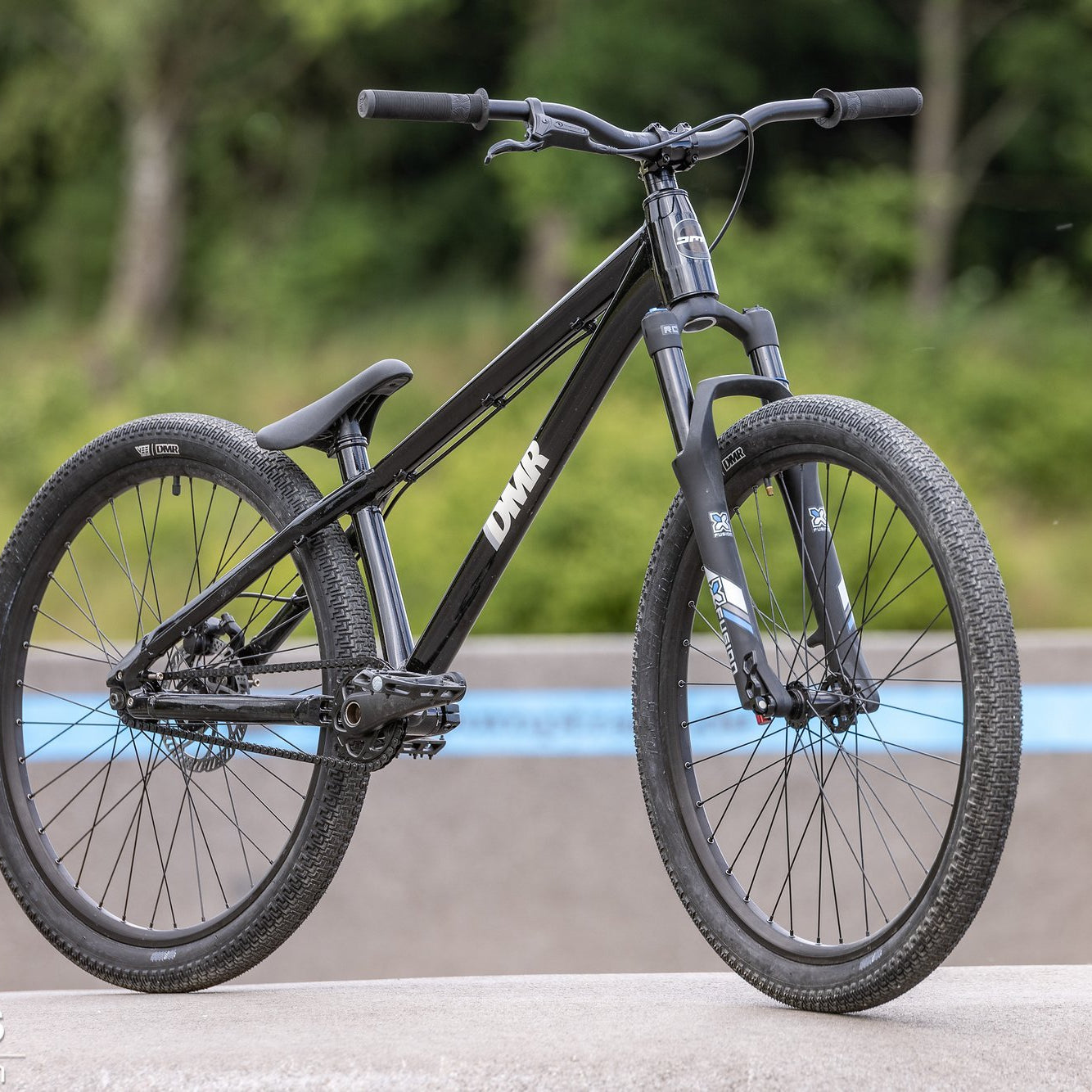 Black DMR Rhythm Dirt Jumping off-road mountain cycling cycle bicycle bike, stood upon a concrete surface, with a woodland background containing trees and bushes.