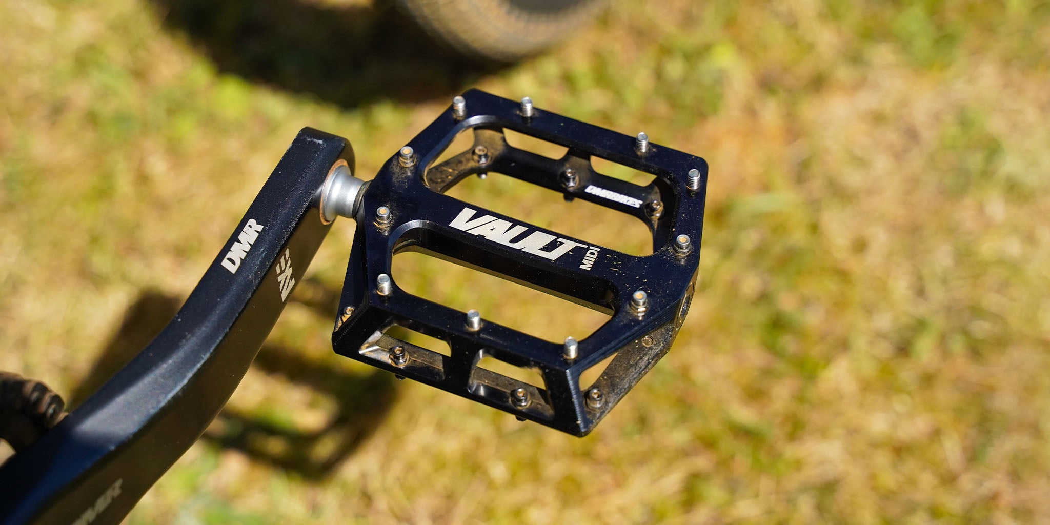 Gloss Black DMR Vault Midi off-road mountain cycling cycle bike bicycle pedal pedalling device, photographed above a grass-covered floor, as seen in the background.
