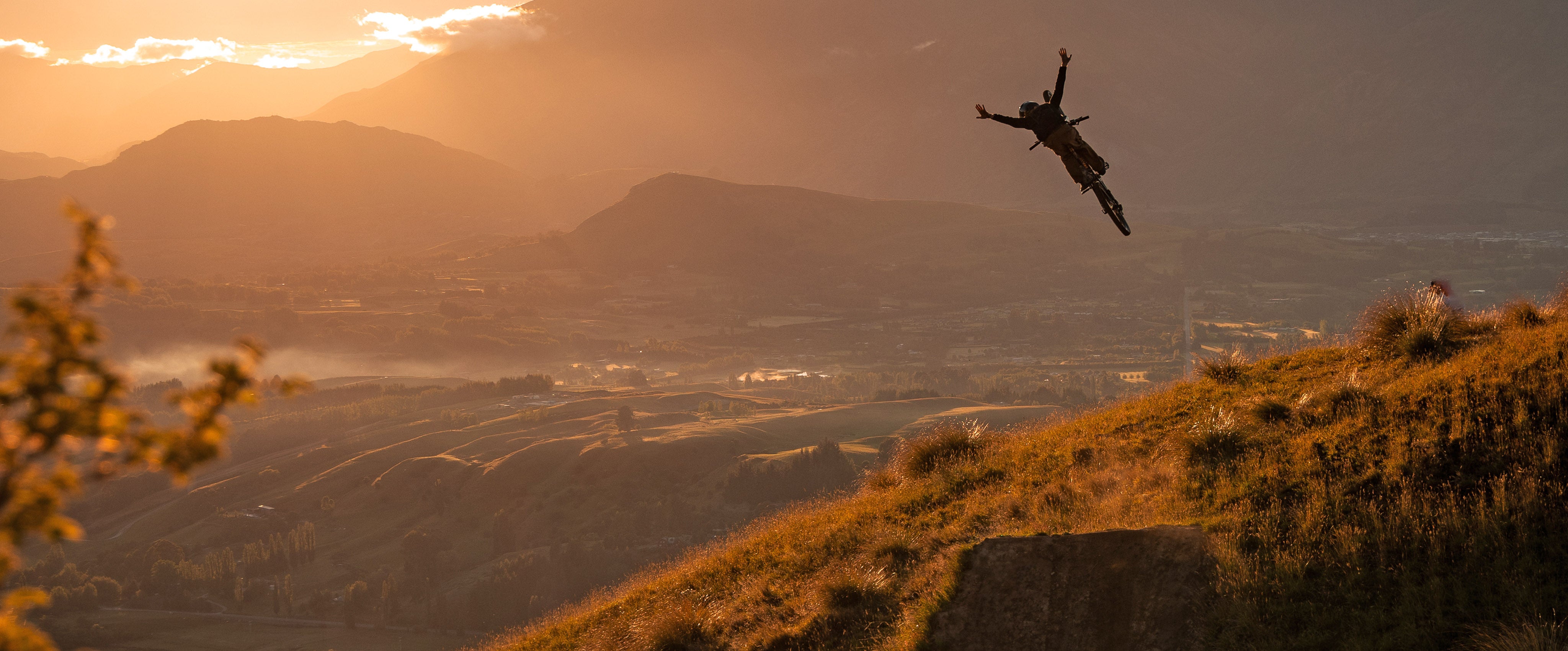 Person on a mountain bike jumping over a hill with a sunset landscape in the background