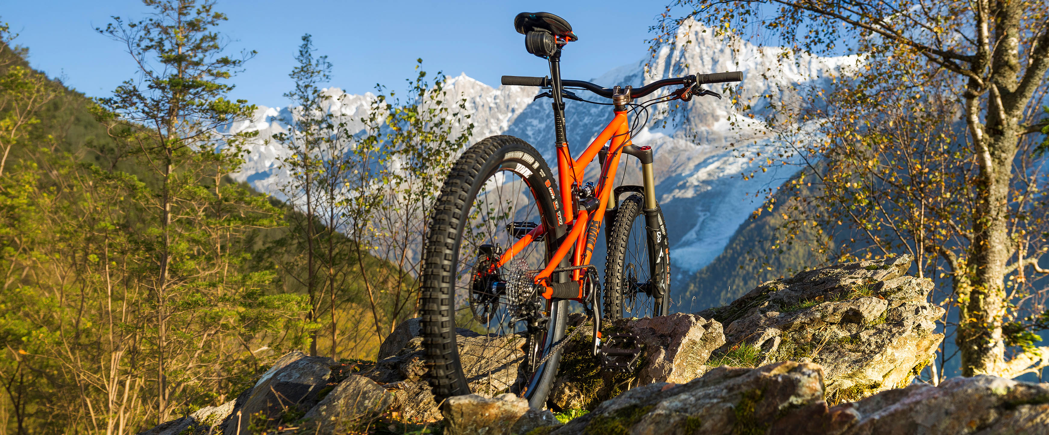 An orange full-suspension mountain bike is parked on rocks in a forest with snow-capped mountains in the background under a blue sky.