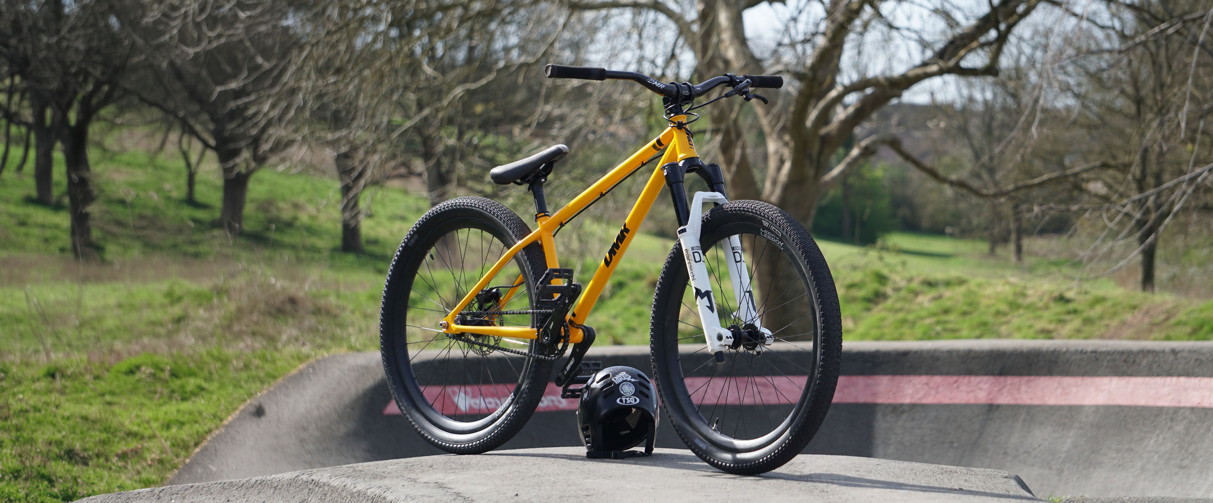 Yellow DMR Sect dirt jump bicycle on a concrete pump track with a helmet nearby, set against a natural background.