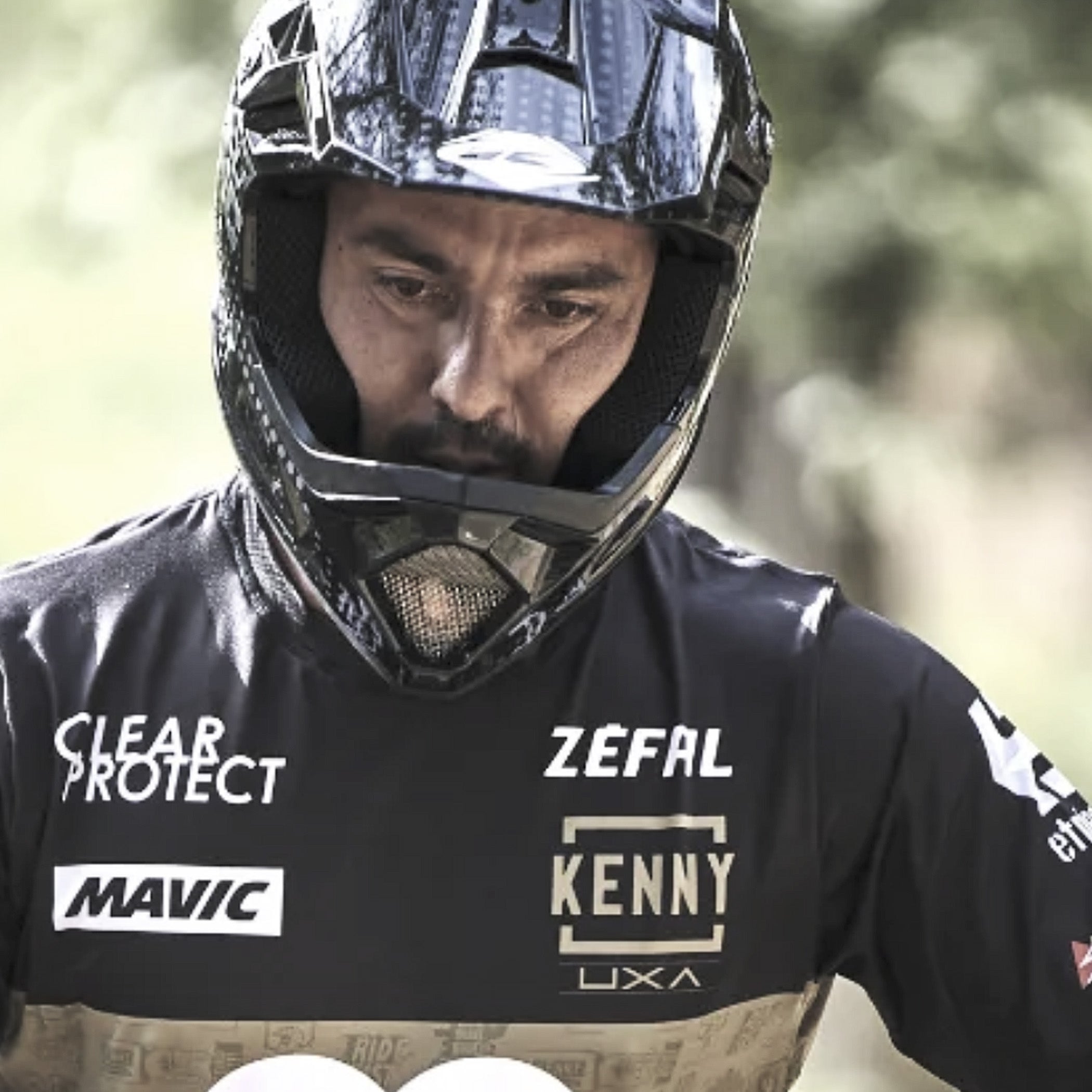 A close-up of a man wearing a black full-face helmet and a black racing jersey with sponsor logos 