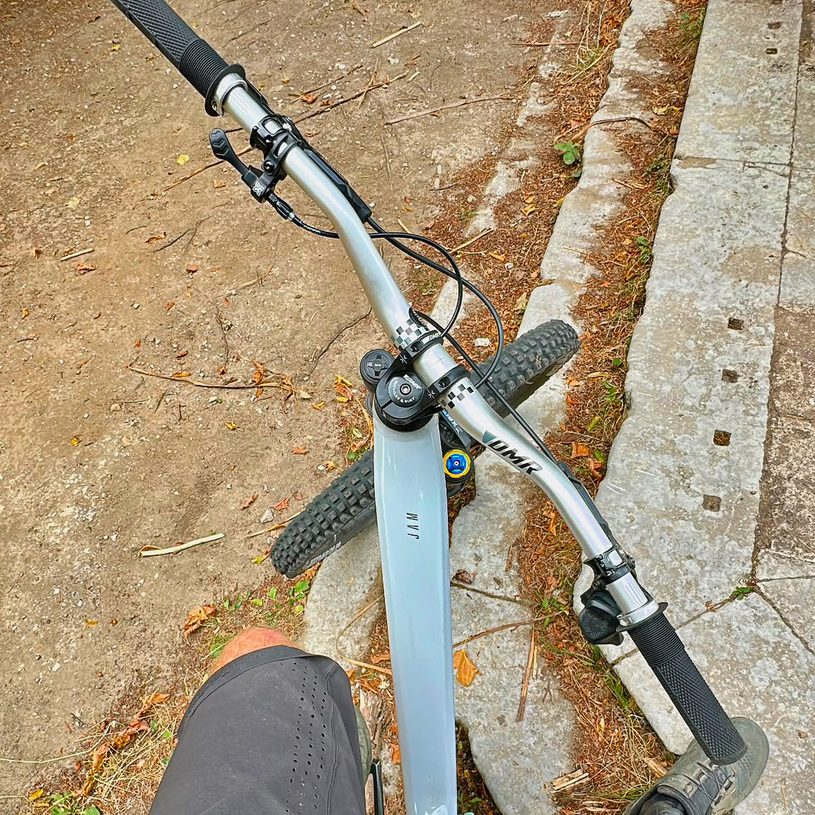 A close-up, top-down view of a mountain bike's handlebars and frame, showing a silver MTB frame with black grips and components, positioned on a dirt path next to a concrete sidewalk.