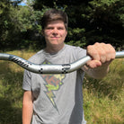 A person outdoors holding a silver mountain bike handlebar with black graphics. The person is wearing a gray graphic t-shirt, and the background shows green trees and grass.