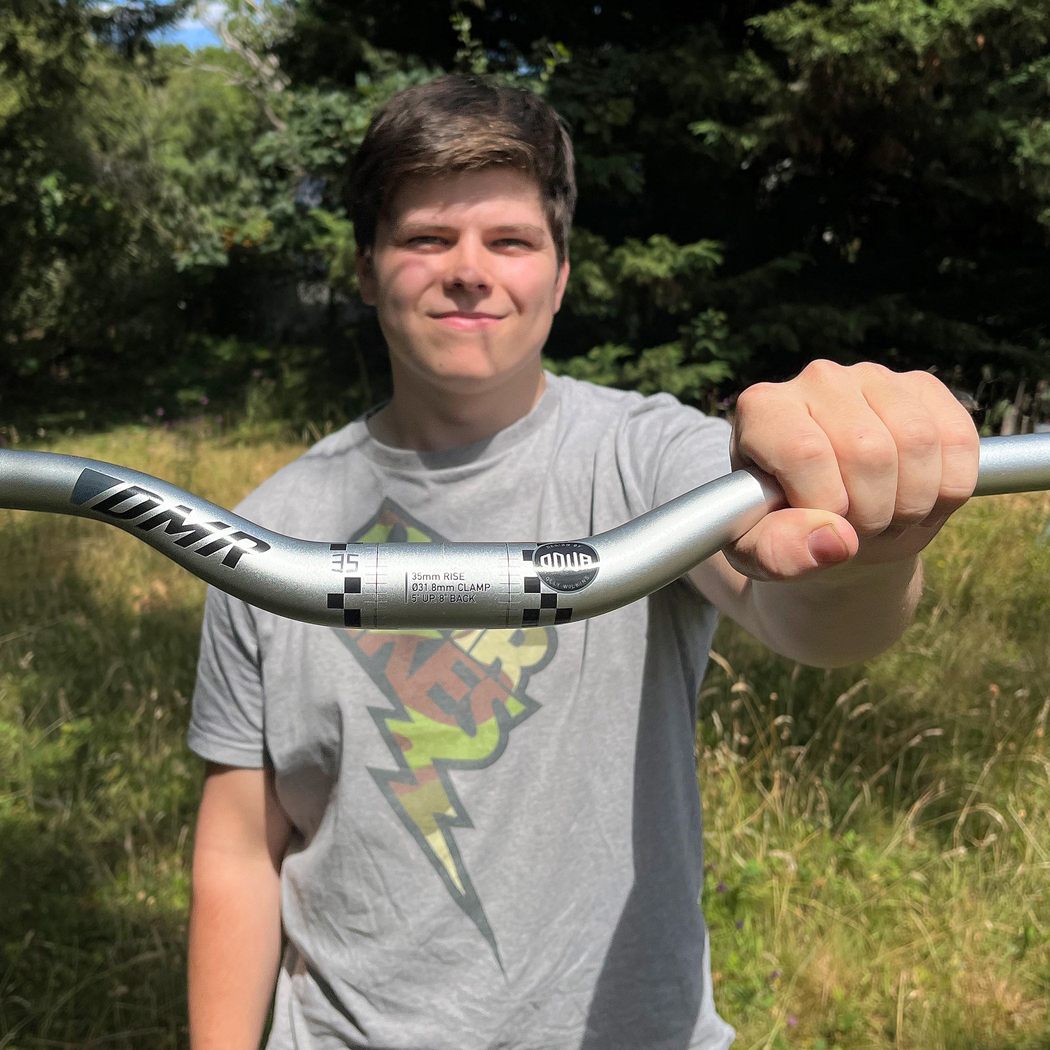 A person outdoors holding a silver mountain bike handlebar with black graphics. The person is wearing a gray graphic t-shirt, and the background shows green trees and grass.