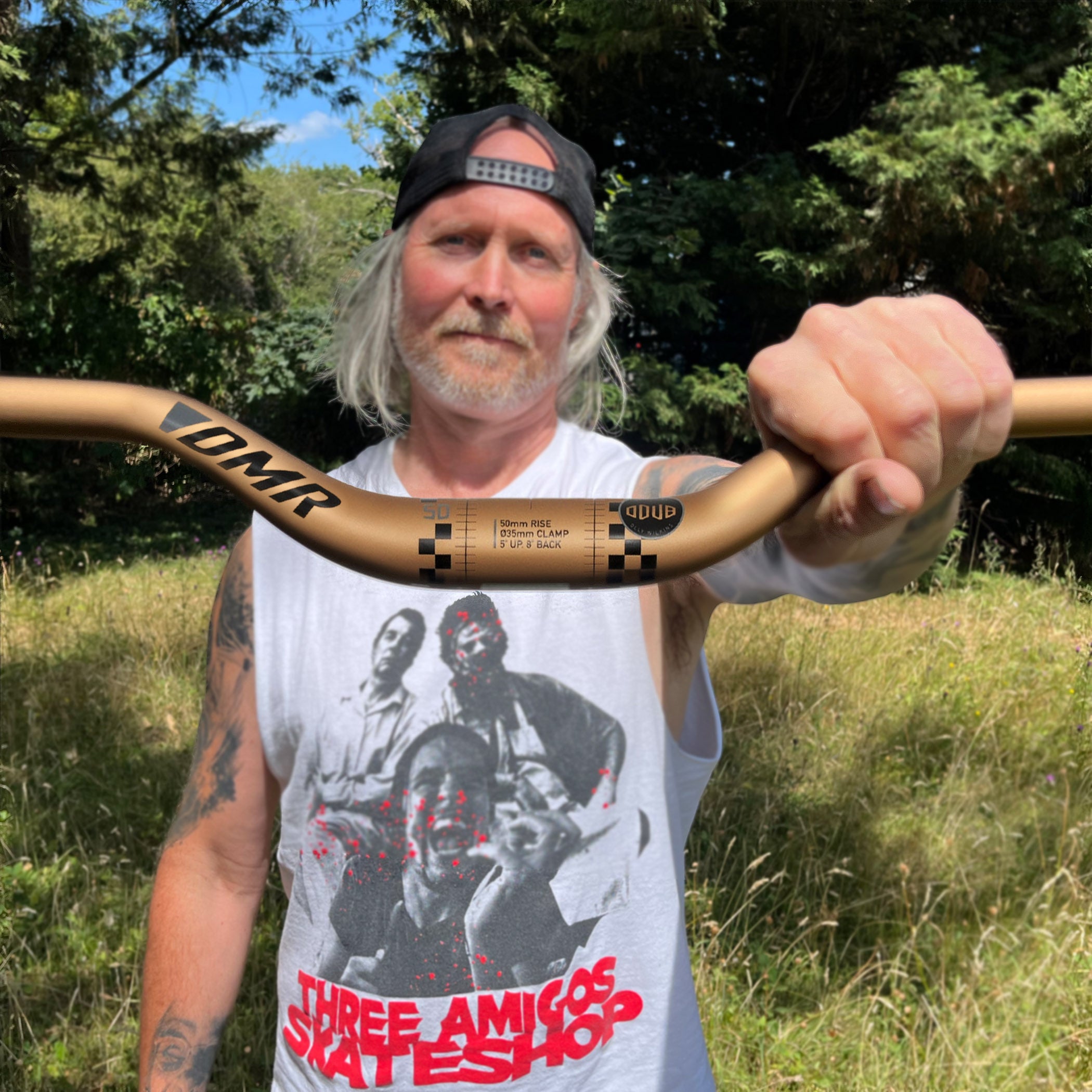 A man outdoors in a grassy field holding a gold-coloured MTB bike handlebar. The handlebar features black branding including 'DMR' and 'ODUB'.