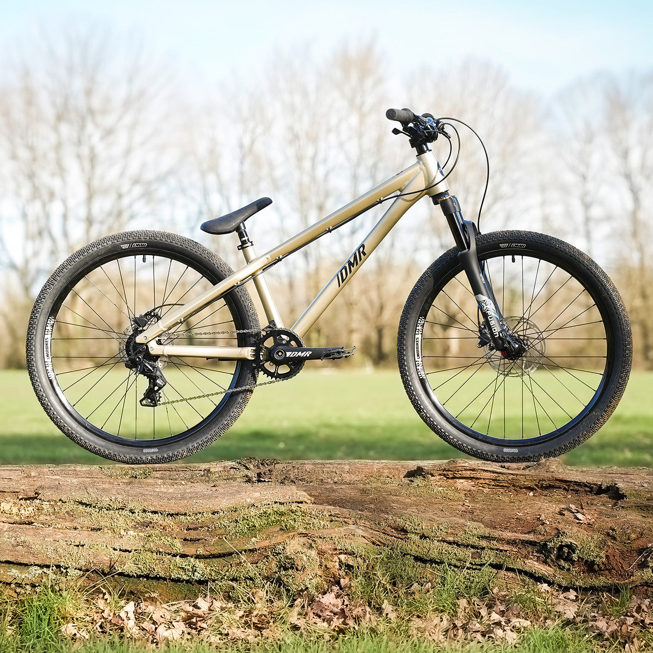 DMR Rhythm Expert bike, in Sola Gold, on a log, with a field and trees in the background.