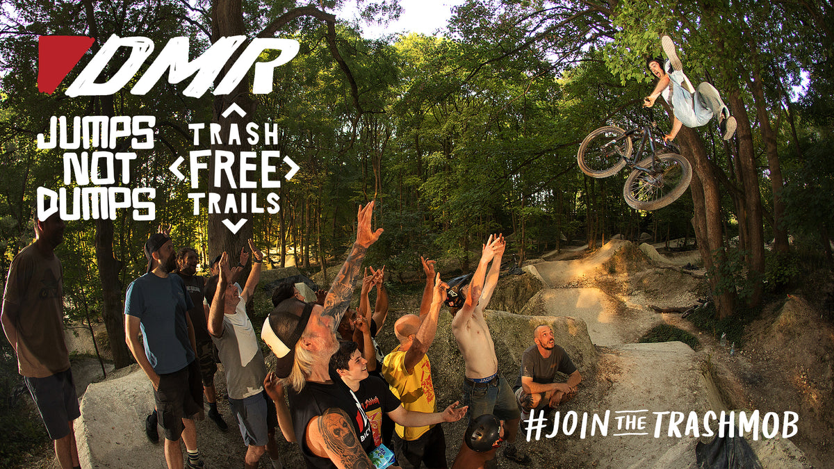 A group of people, the DMR team, gathered at a forest mountain bike jump watching a rider perform a high aerial trick. Large overlaid text reads 'DMR', 'JUMPS NOT DUMPS', 'TRASH FREE TRAILS', and '#JOIN THE TRASHMOB'.