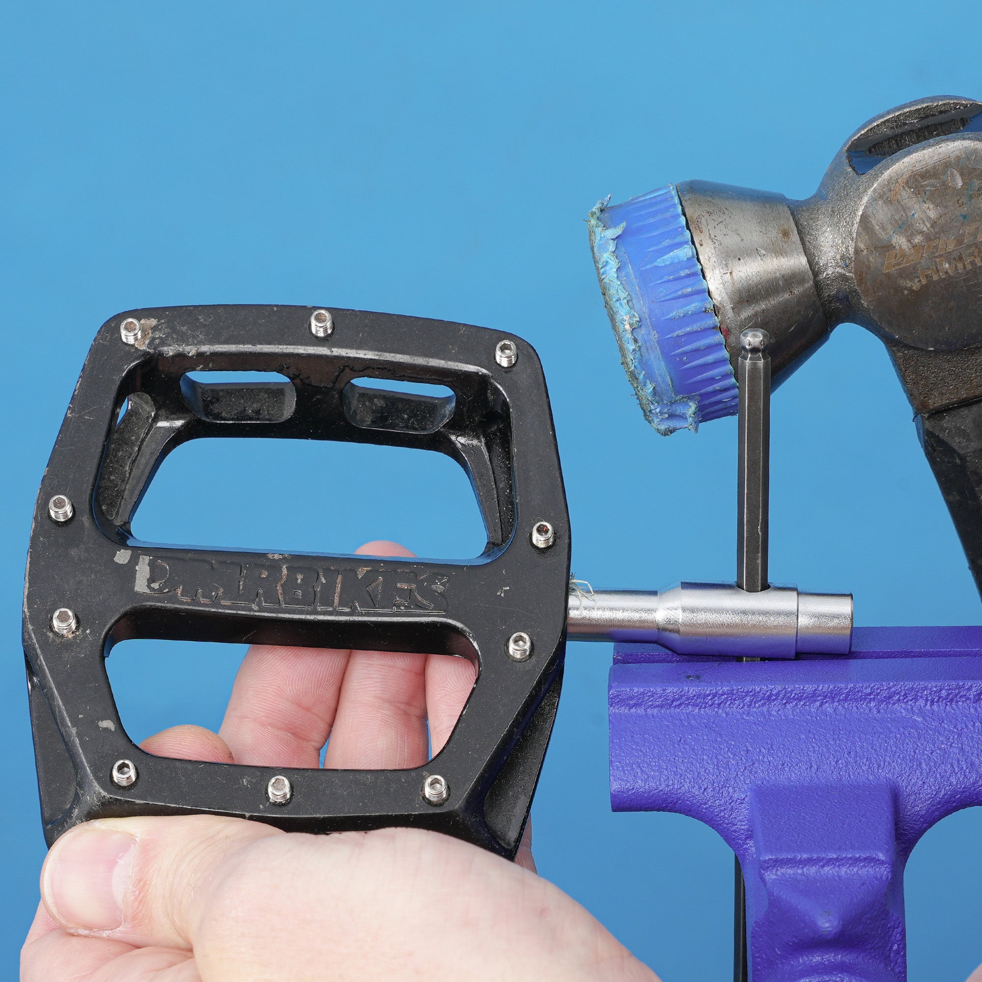 silver dmr bikes bicycles vmax vault mag magnesium midi v12 v11 flat4 flat 4 four versa clip clipless pedals pedal service servicing tool in use with allen key and hammer in a vice on a blue background