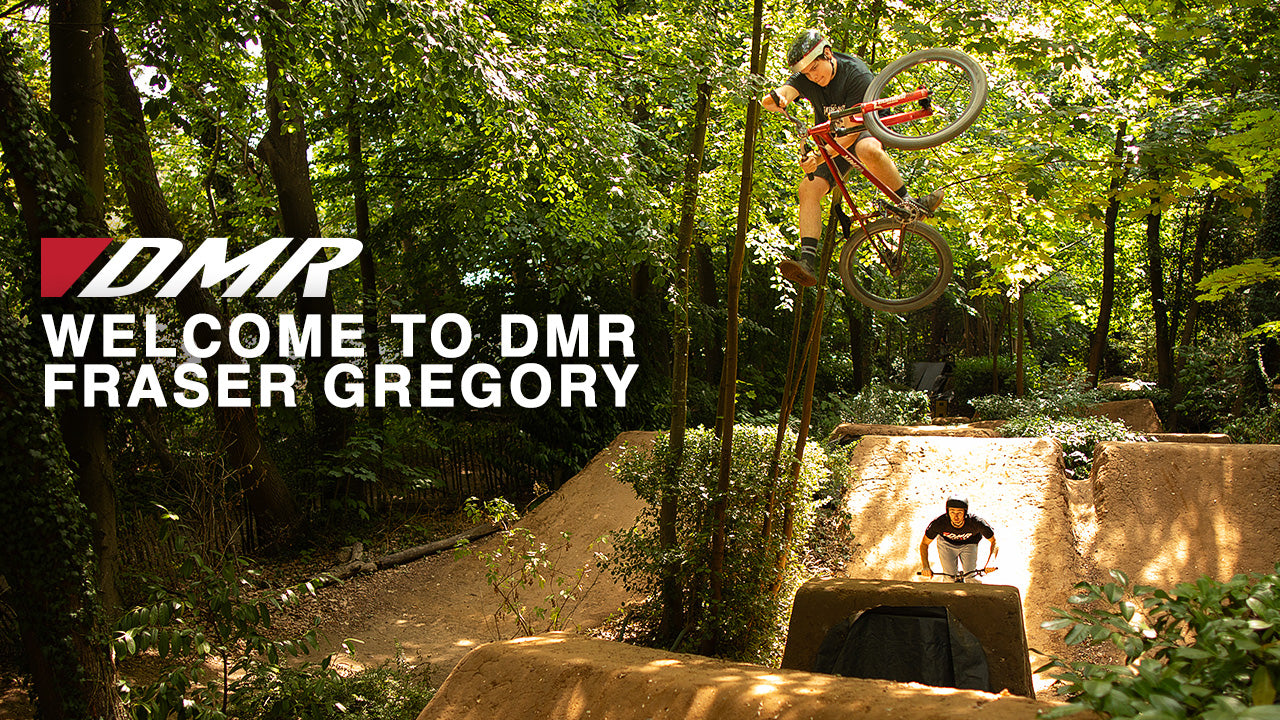 A mountain biker mid-air on a DJ-style bike above a dirt jump in a wooded outdoor setting, with another rider below. Large text on the left reads 'DMR WELCOME TO DMR FRASER GREGORY'.
