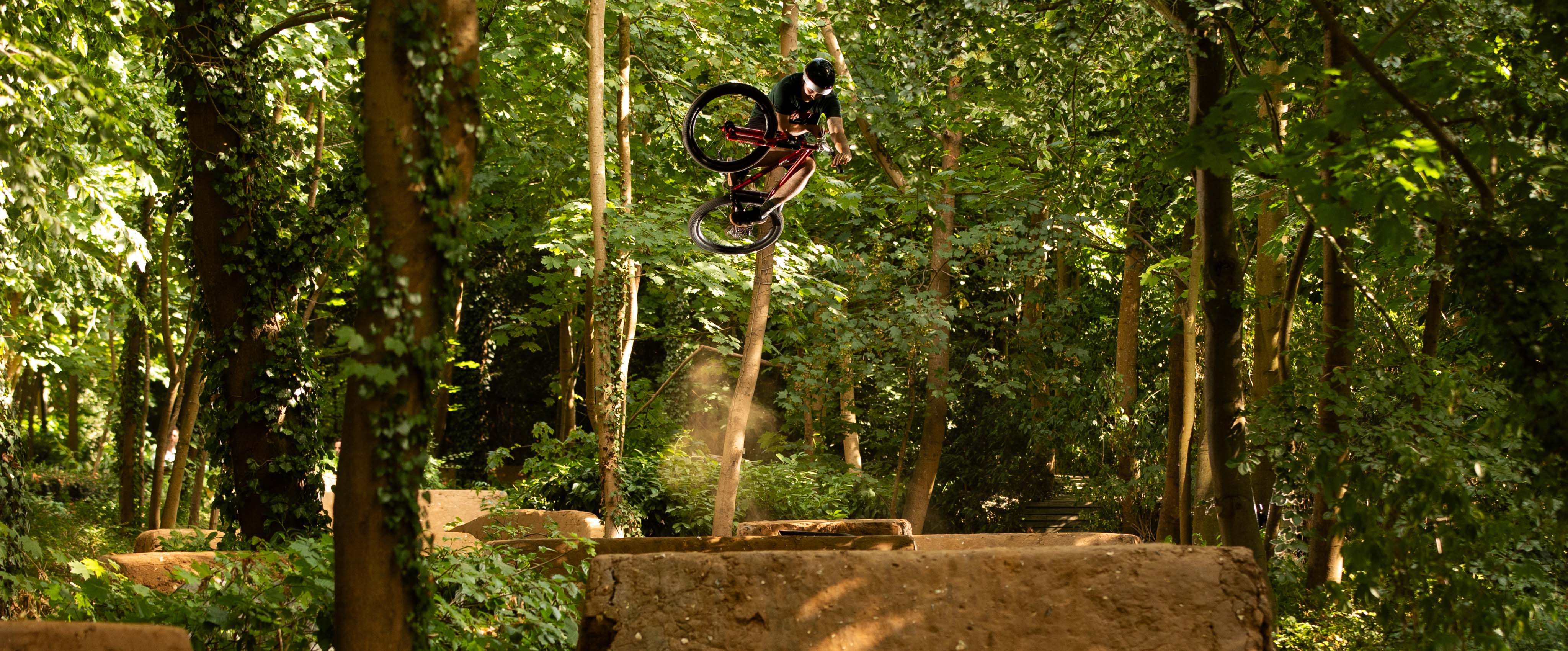A mountain biker mid-air performing a jump in a dense green forest, with dirt ramps visible below.