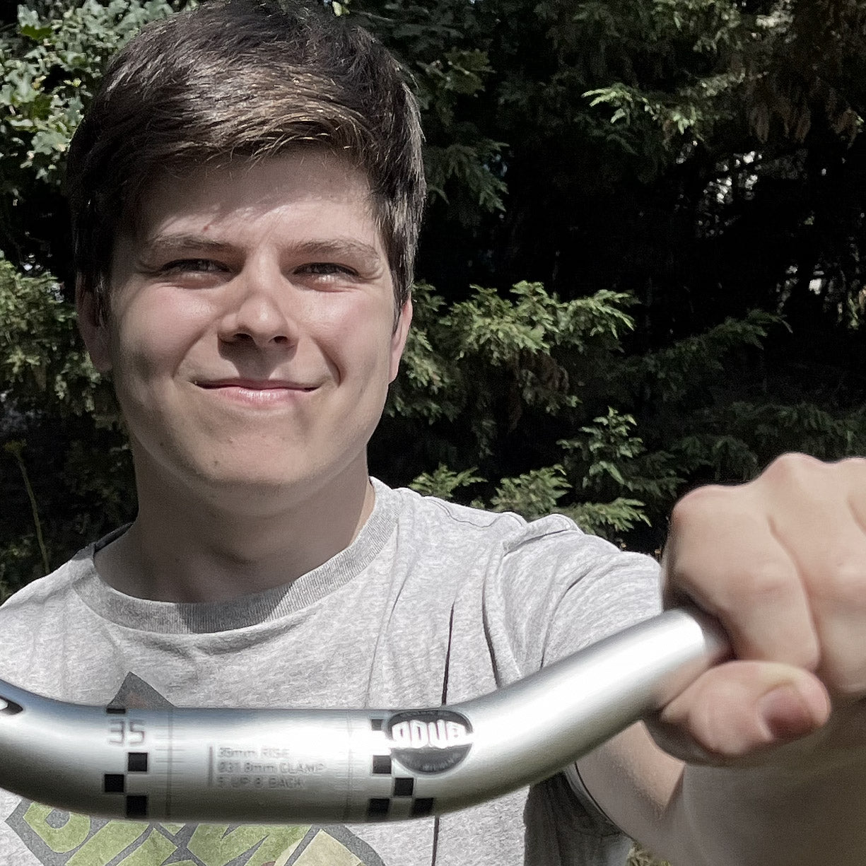 A smiling young man outdoors holding a silver mountain bike handlebar. The handlebar features the 'ODUB' brand logo, and the background consists of green trees.