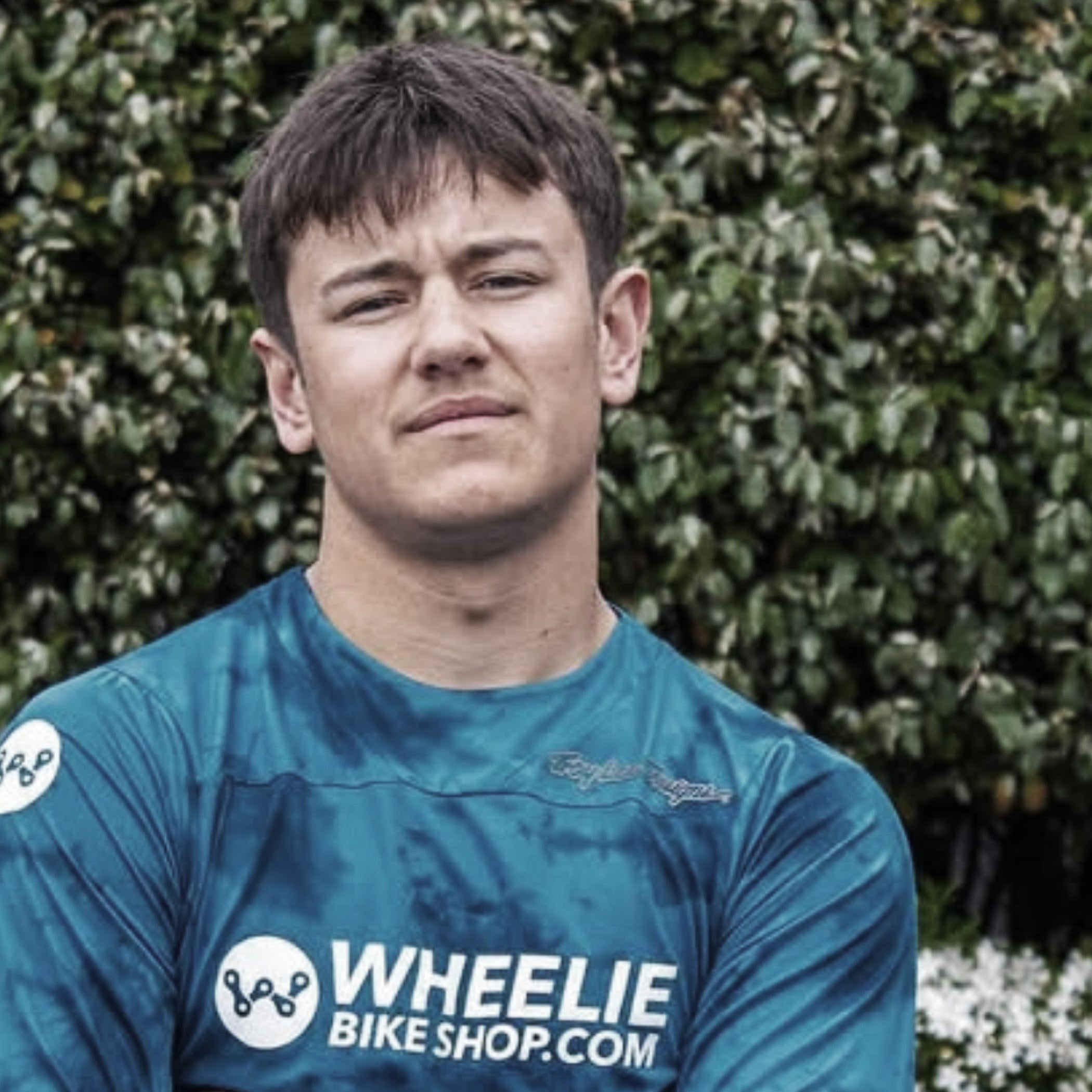 Person wearing a blue t-shirt with 'Wheelie Bike Shop.com' logo in front of a green hedge.