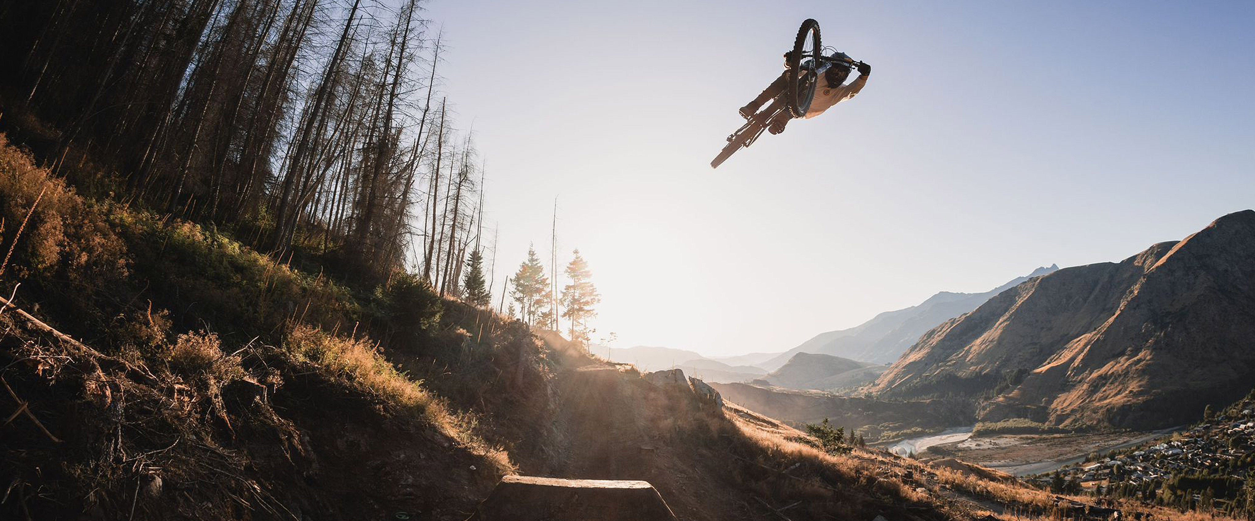 Biker in mid-air over a dirt trail with mountains and trees in the background