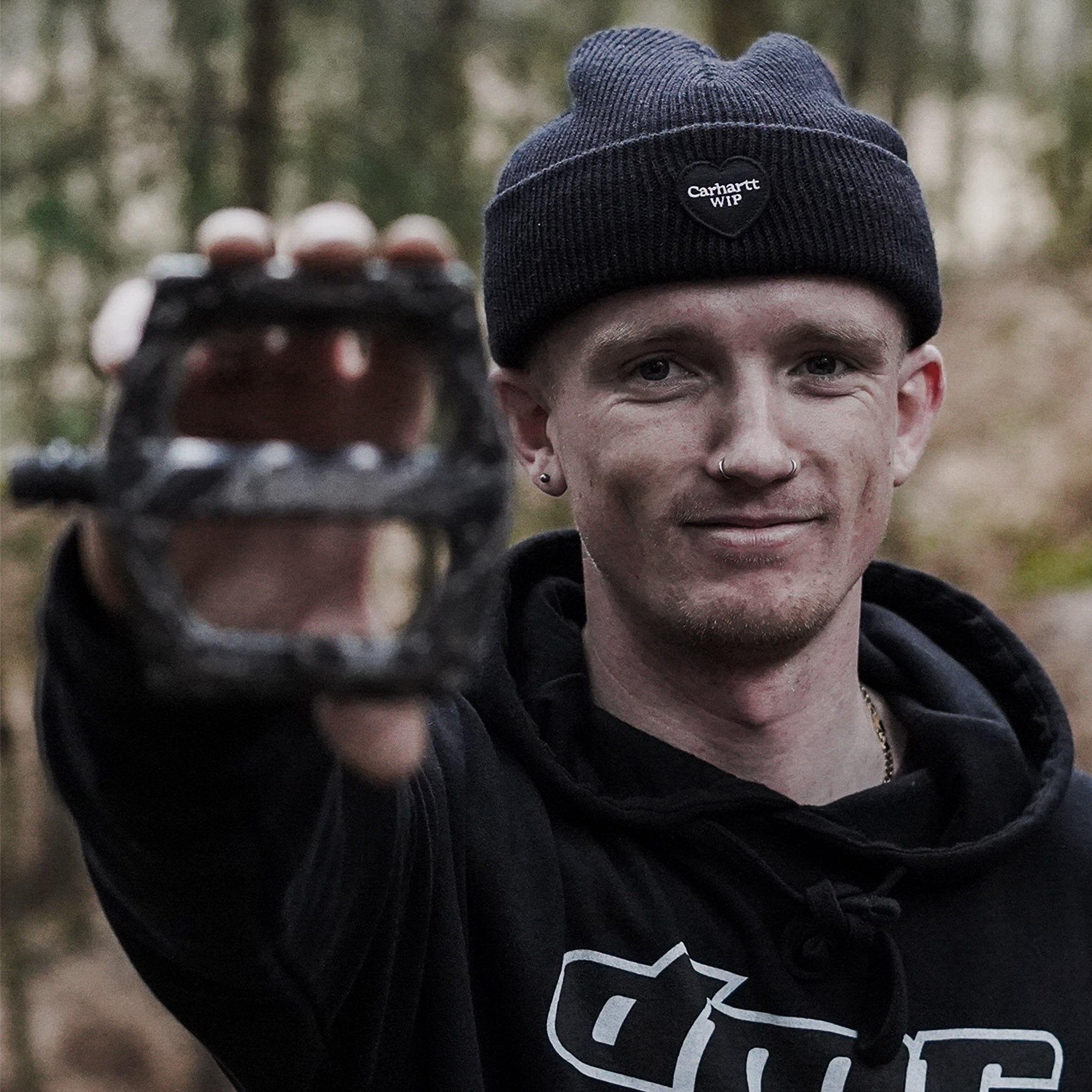 Person wearing a black hoodie and navy beanie with a logo, holding a metal object outdoors.