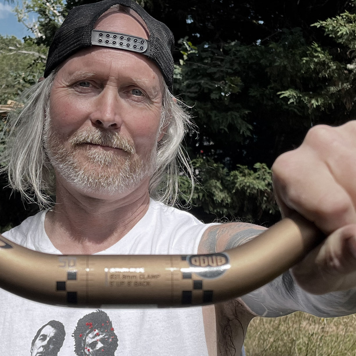 A close-up lifestyle shot of a man with long gray hair and a beard, wearing a black baseball cap backwards and a white graphic t-shirt. He is holding a gold-colored bicycle handlebar with 'ODUB' branding in an outdoor setting with green trees in the background.
