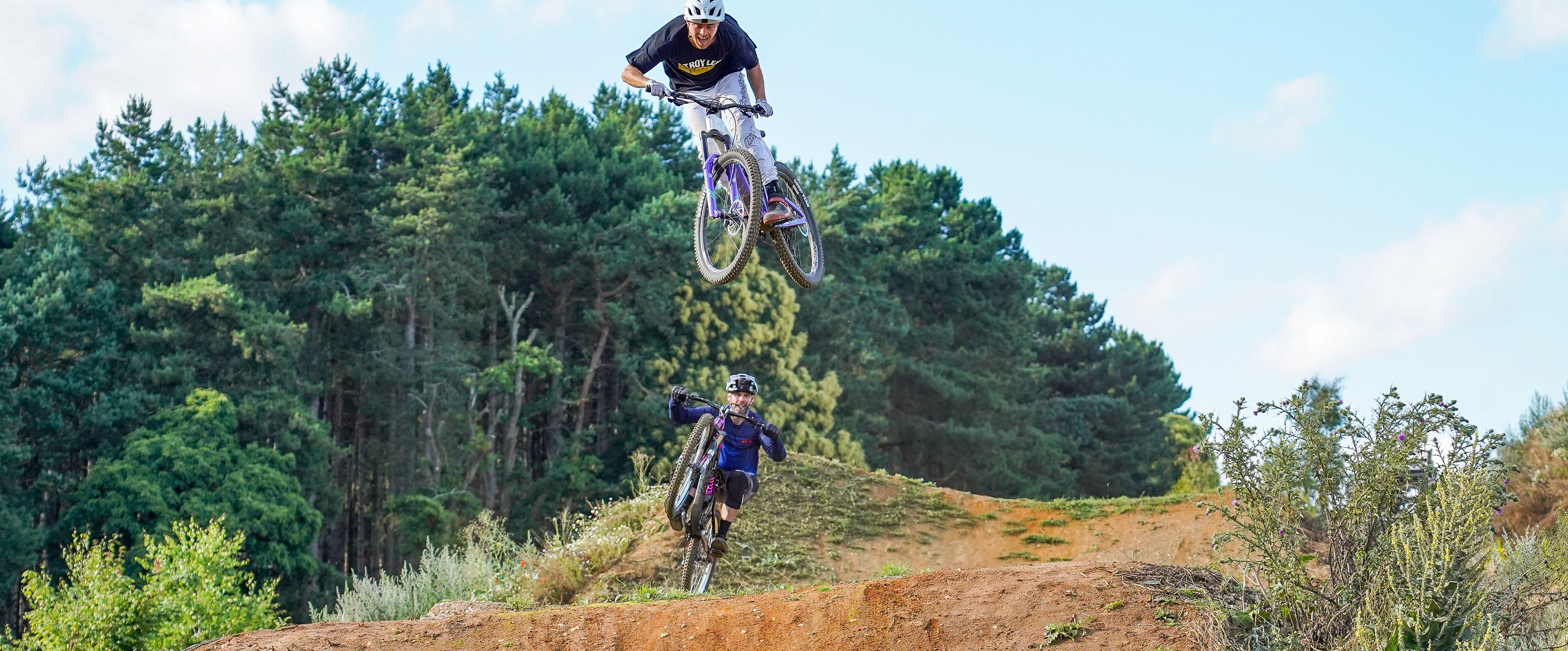 Two people on mountain bikes performing a jump in a forested area.