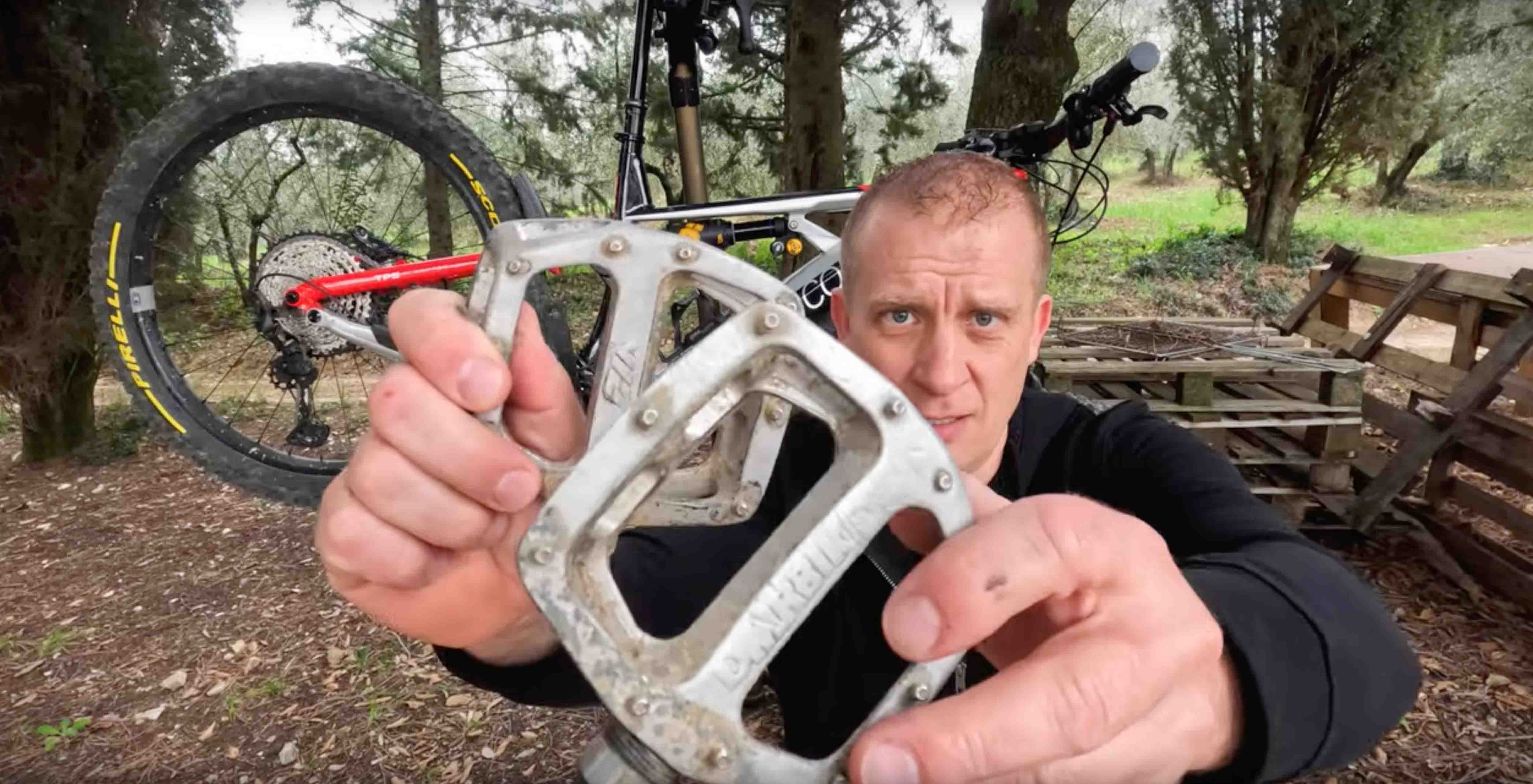 A man is holding a DMR V12 pedal pedalling product, designed for off-road mountain cycling. This DMR V12 pedal pedalling product, designed for off-road mountain cycling, is silver. In the background, is an off-road mountain cycling bicycle, in a bicycle-holding apparatus, with trees and a wooden structure.