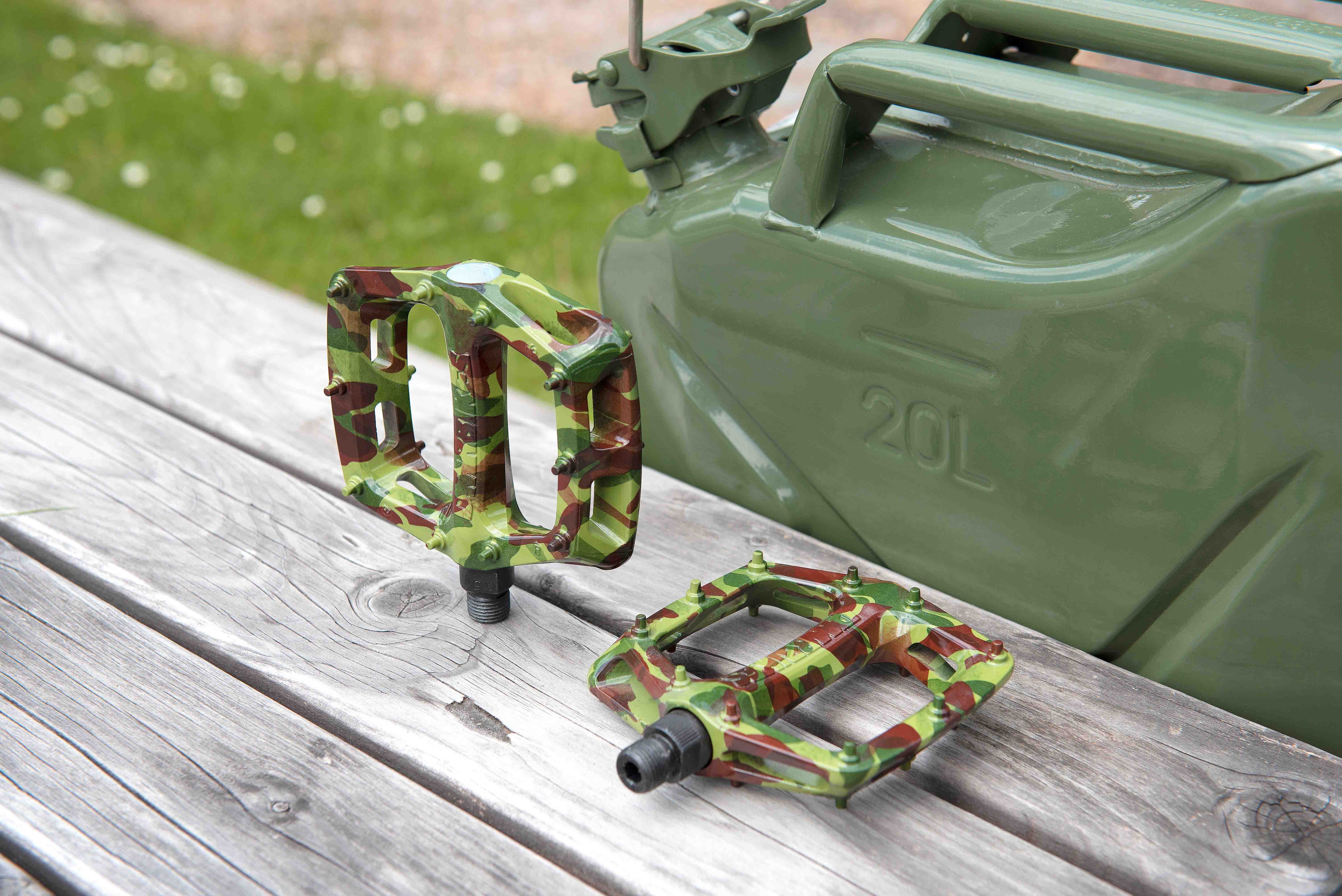 DMR V6 plastic off-road mountain cycling pedal pedalling products, situated on top of a wooden bench table top surface. These DMR V6 plastic off-road mountain cycling pedal pedalling products have a green and brown coloured camouflage pattern. There is a large green receptacle in the background.
