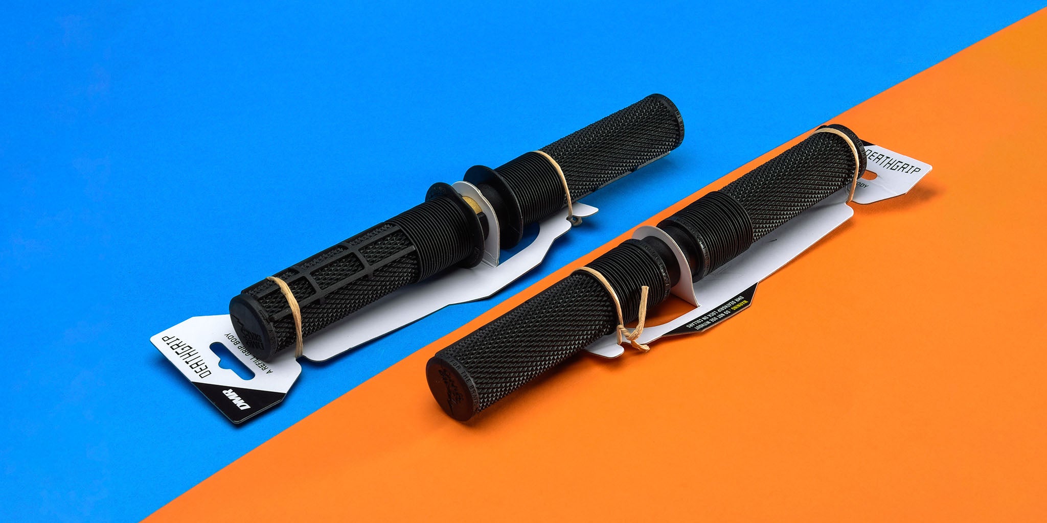 Two DMR Deathgrip off-road mountain cycling hand hold grip refill grip refillable grips, one on a blue background, the other on orange. These DMR Deathgrip off-road mountain cycling hand hold grip refill grip refillable grips are Black in colour, and contained within their packaging, which is White.
