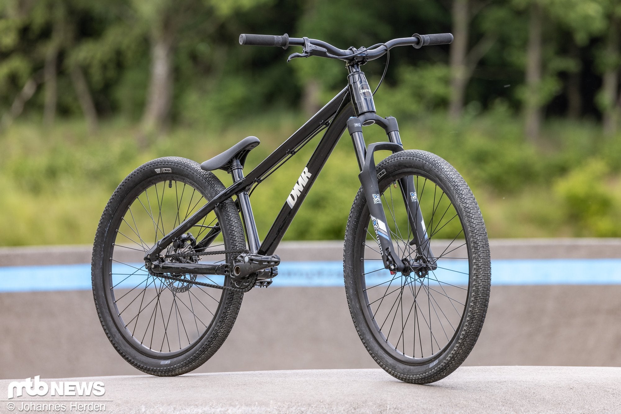 Black DMR Rhythm Dirt Jumping off-road mountain cycling cycle bicycle bike, stood upon a concrete surface, with a woodland background containing trees and bushes.