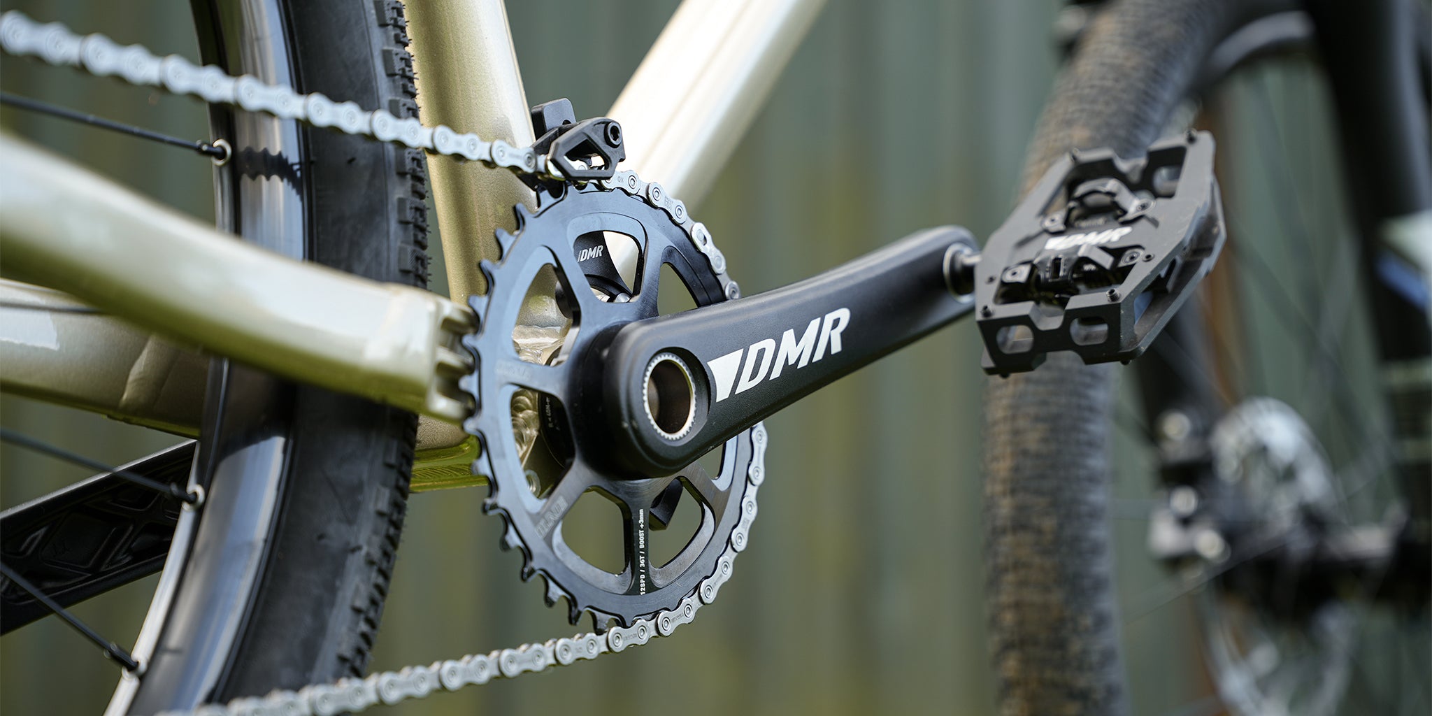 DMR Sect Cranks in Black, fitted to a DMR Rhythm Expert bike, with a blurry green fence in the background.