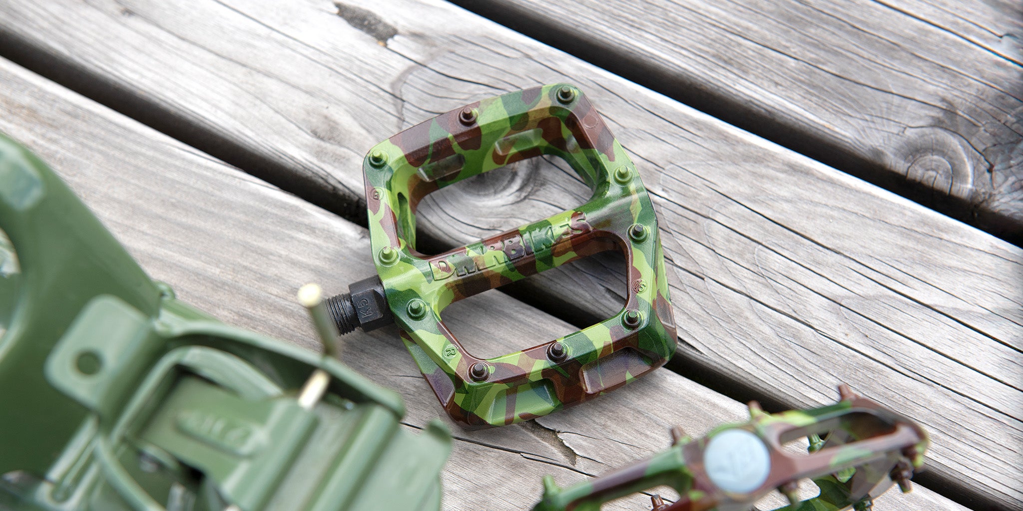 DMR V6 SE Special Edition off-road mountain cycling pedal pedalling product, placed upon a wooden table top. The DMR V6 SE Special Edition off-road mountain cycling pedal pedalling product is coloured with a green and brown camouflage pattern.