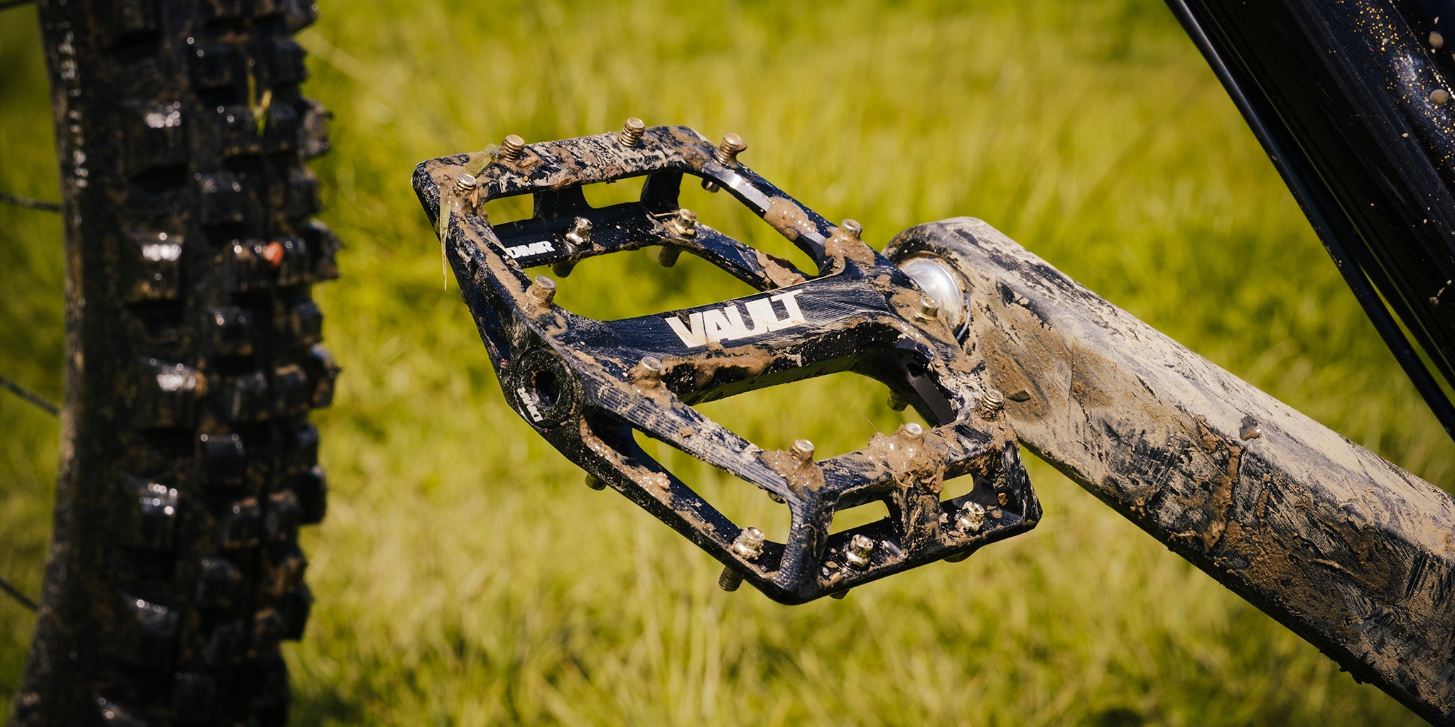 A DMR Vault off-road mountain cycling pedal pedalling product, covered in a mud/clay substance. This DMR Vault off-road mountain cycling pedal pedalling product is Black in colour. In the background, there is grass, which is green.